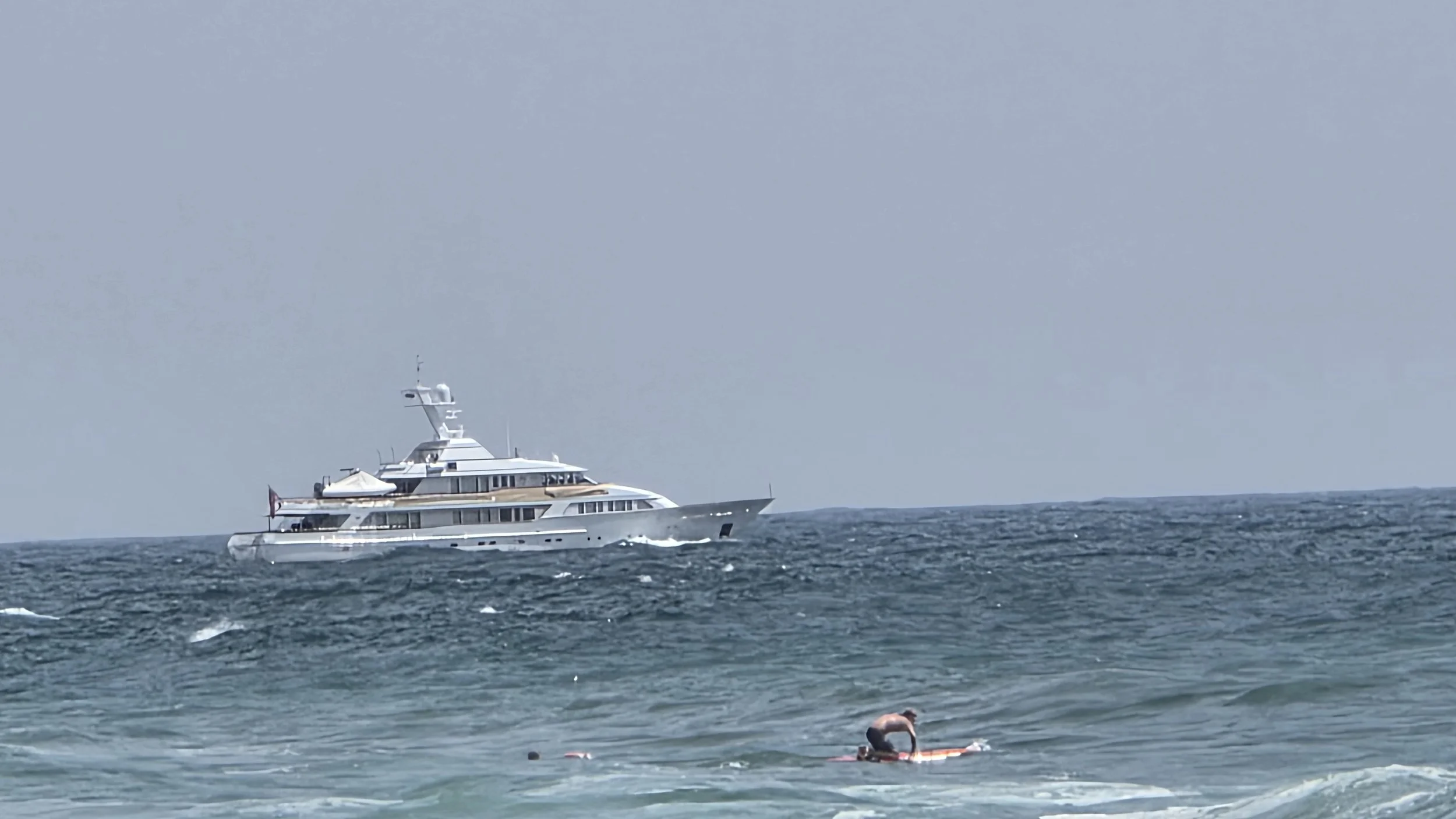 A large yacht sailing in the ocean with waves in the foreground and a person on a surfboard in the water.