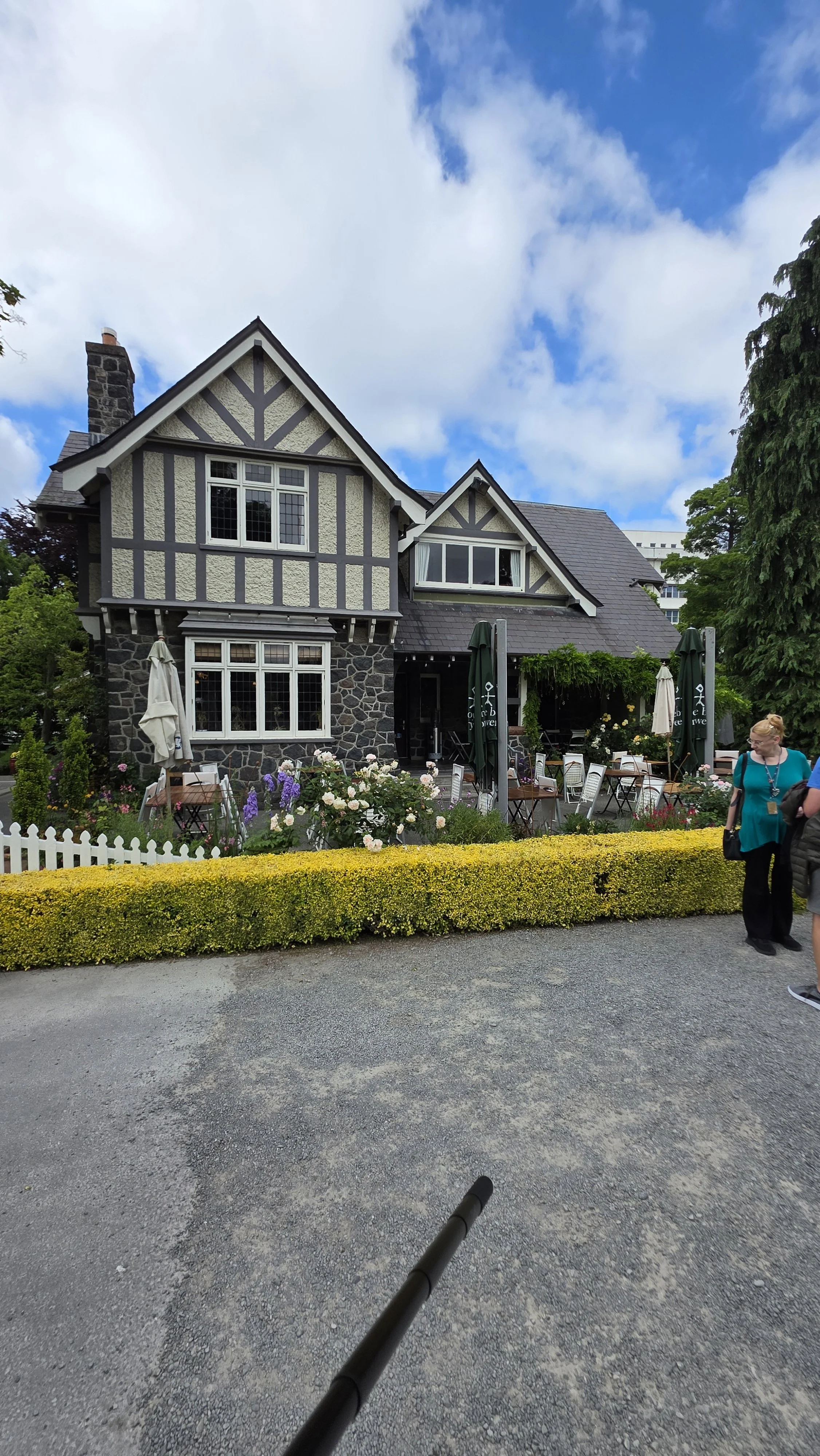A charming, black and white Tudor-style house with a flower garden in front and outdoor seating, under a partly cloudy sky with two people standing on the sidewalk.