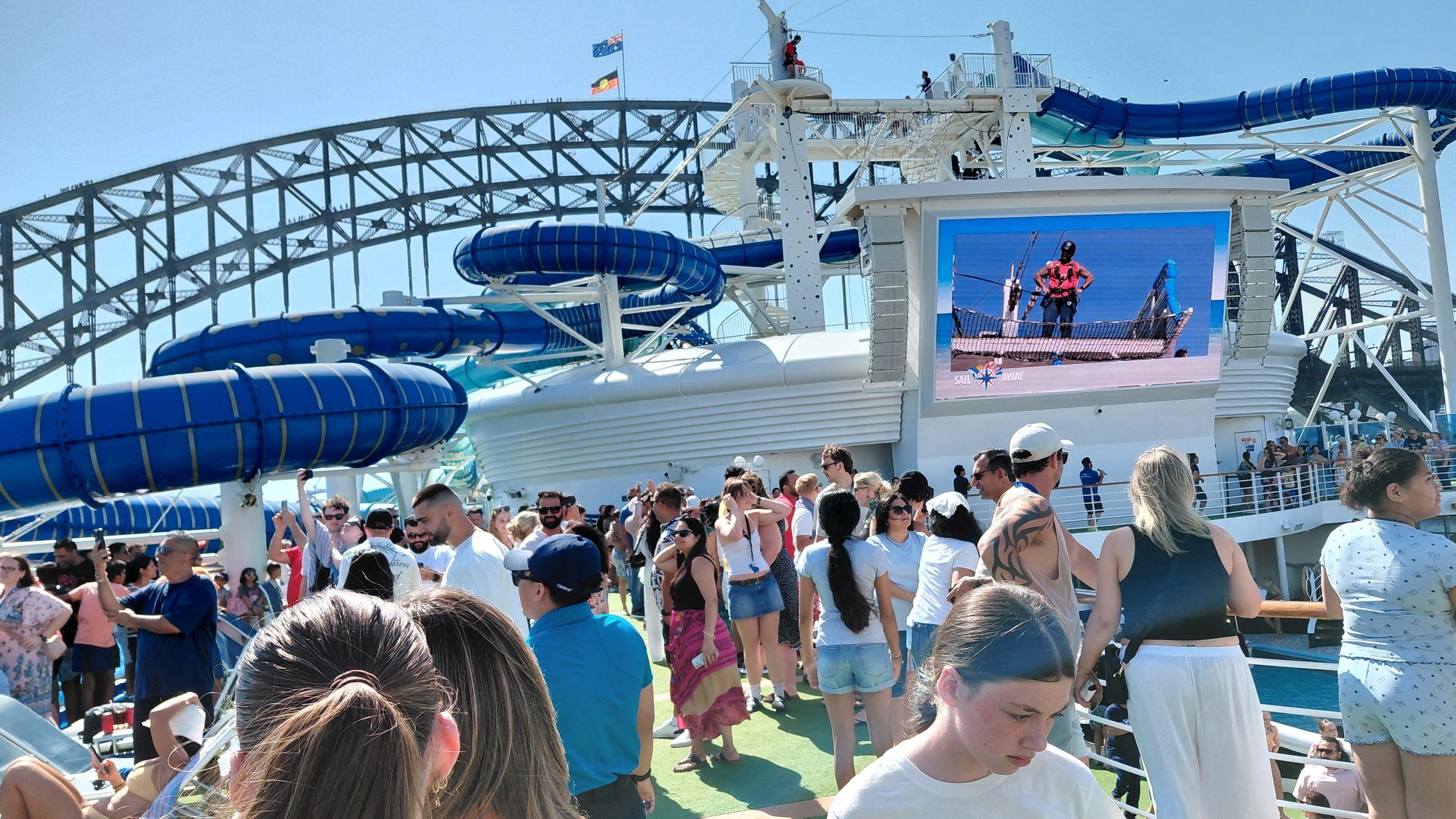 Crowd of people on a cruise ship deck with a waterslide and a large screen displaying an image of a person on a boat