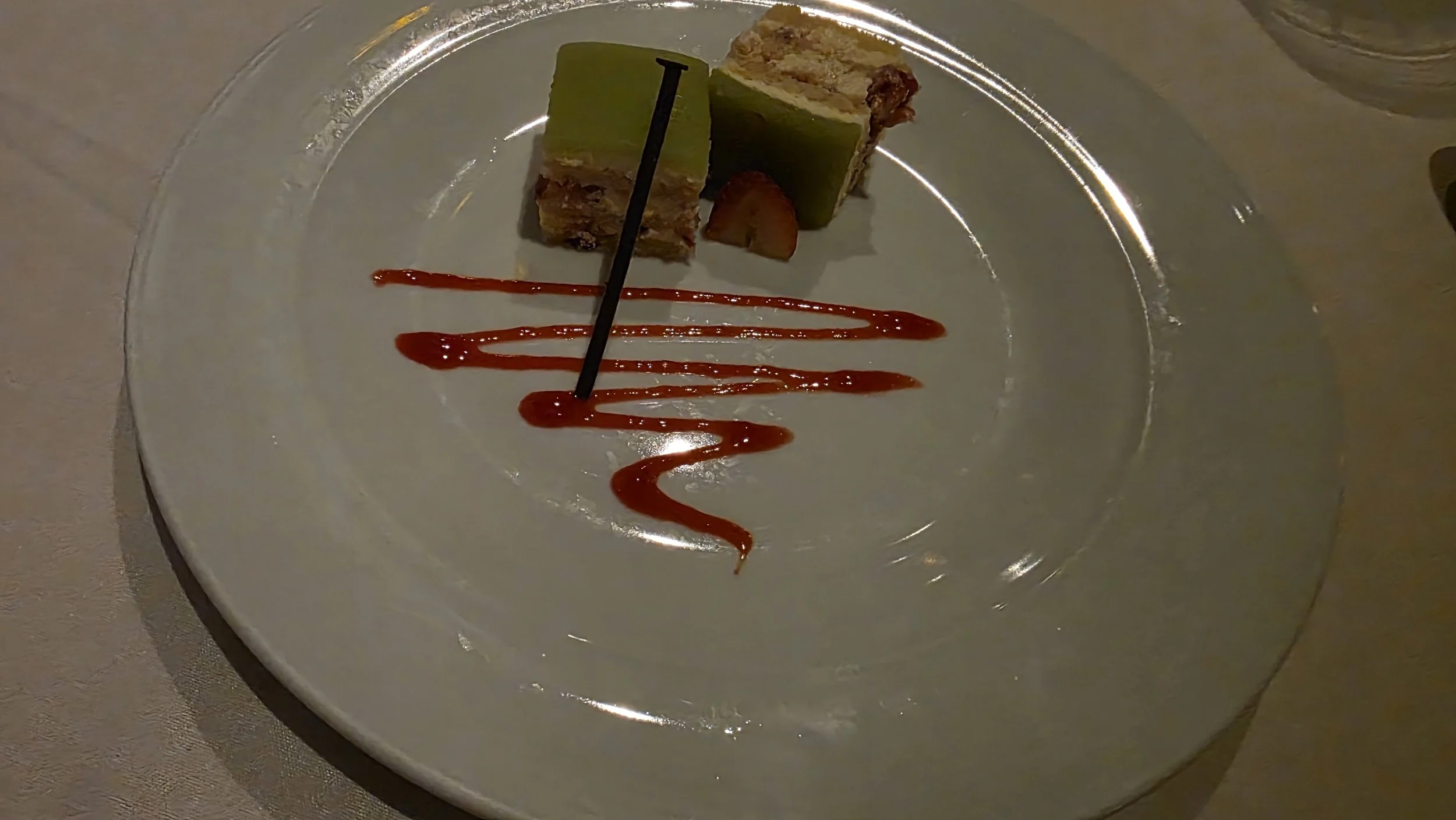 A dessert consisting of stacked layers with green, yellow, and brown colors on a white plate. The dessert is decorated with a zigzag line of red sauce or syrup, and there is a thin black stick inserted into the dessert.