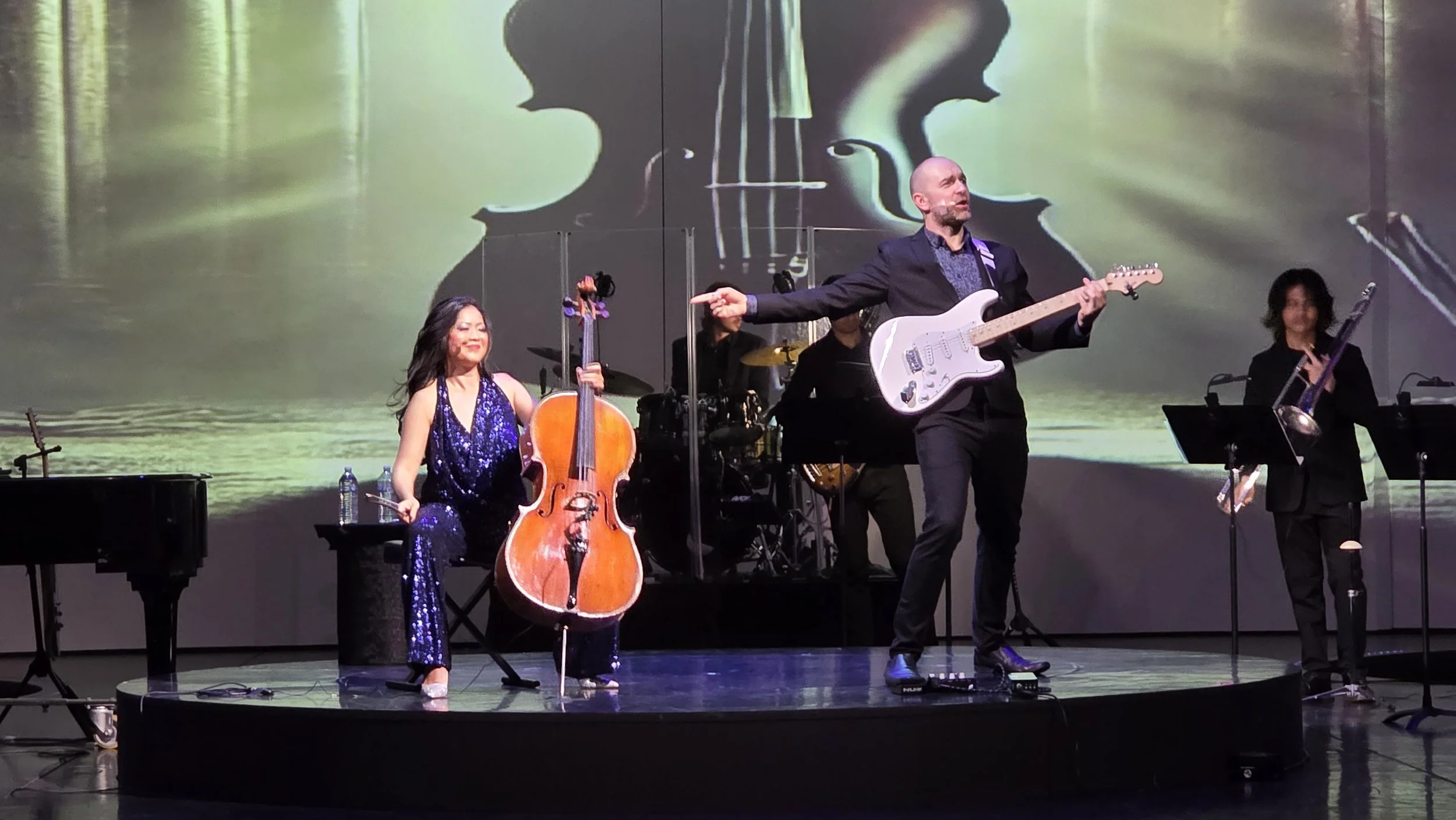 Musicians performing on stage, including a woman in a sparkly blue dress playing a cello, a man in a black suit playing an electric guitar, and other musicians in black playing instruments. There is a large screen behind them displaying a violin silh
