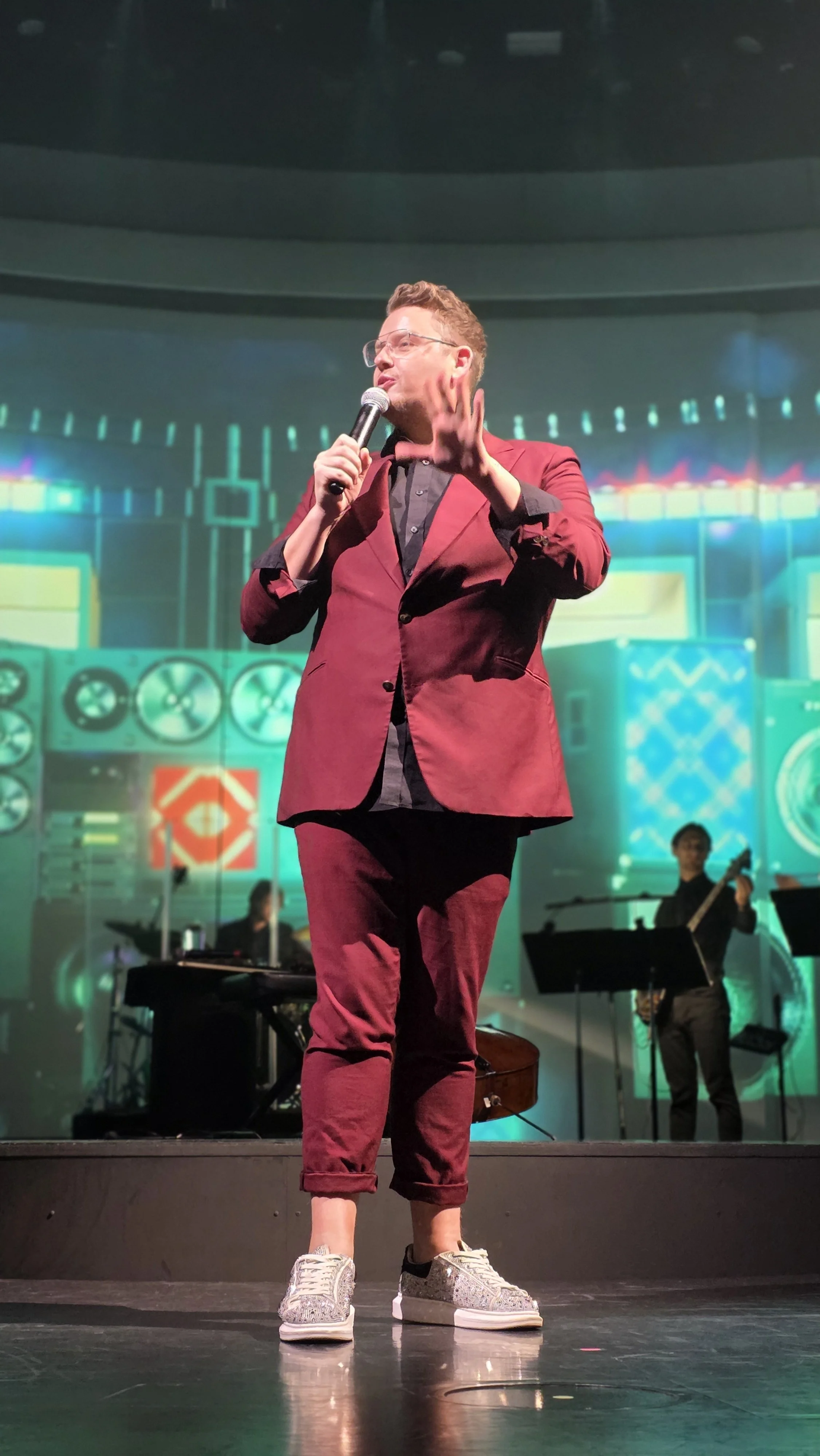 A man in a burgundy suit and glasses is holding a microphone on stage, with a colorful background and a person playing guitar behind him.