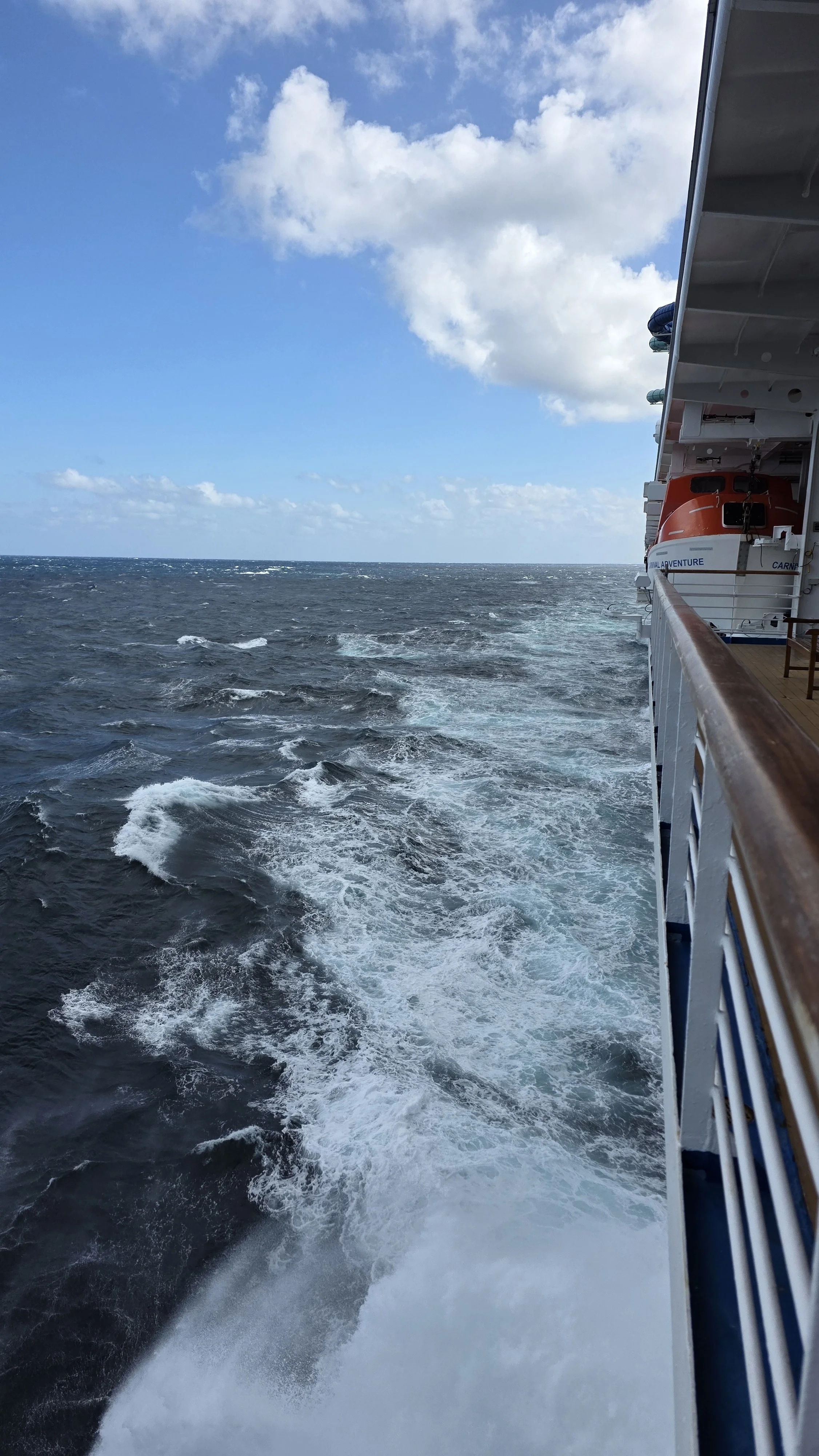 View of ocean water with waves and sky with clouds taken from the side of a cruise ship, showing the ship's railing and part of another lifeboat.