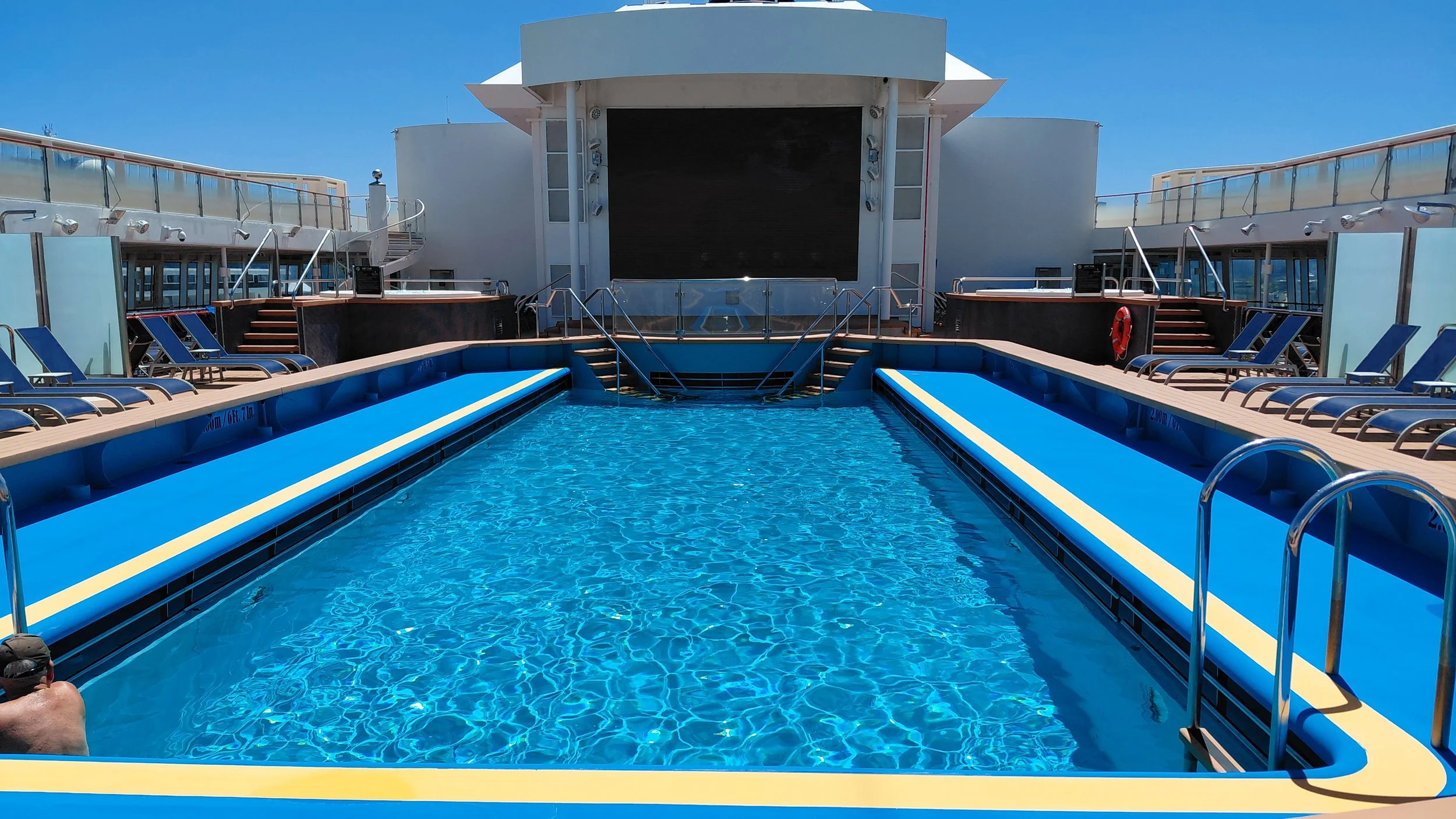 Empty swimming pool area on cruise ship with lounge chairs along sides, stairs leading to upper deck, and a large outdoor movie screen at the back.
