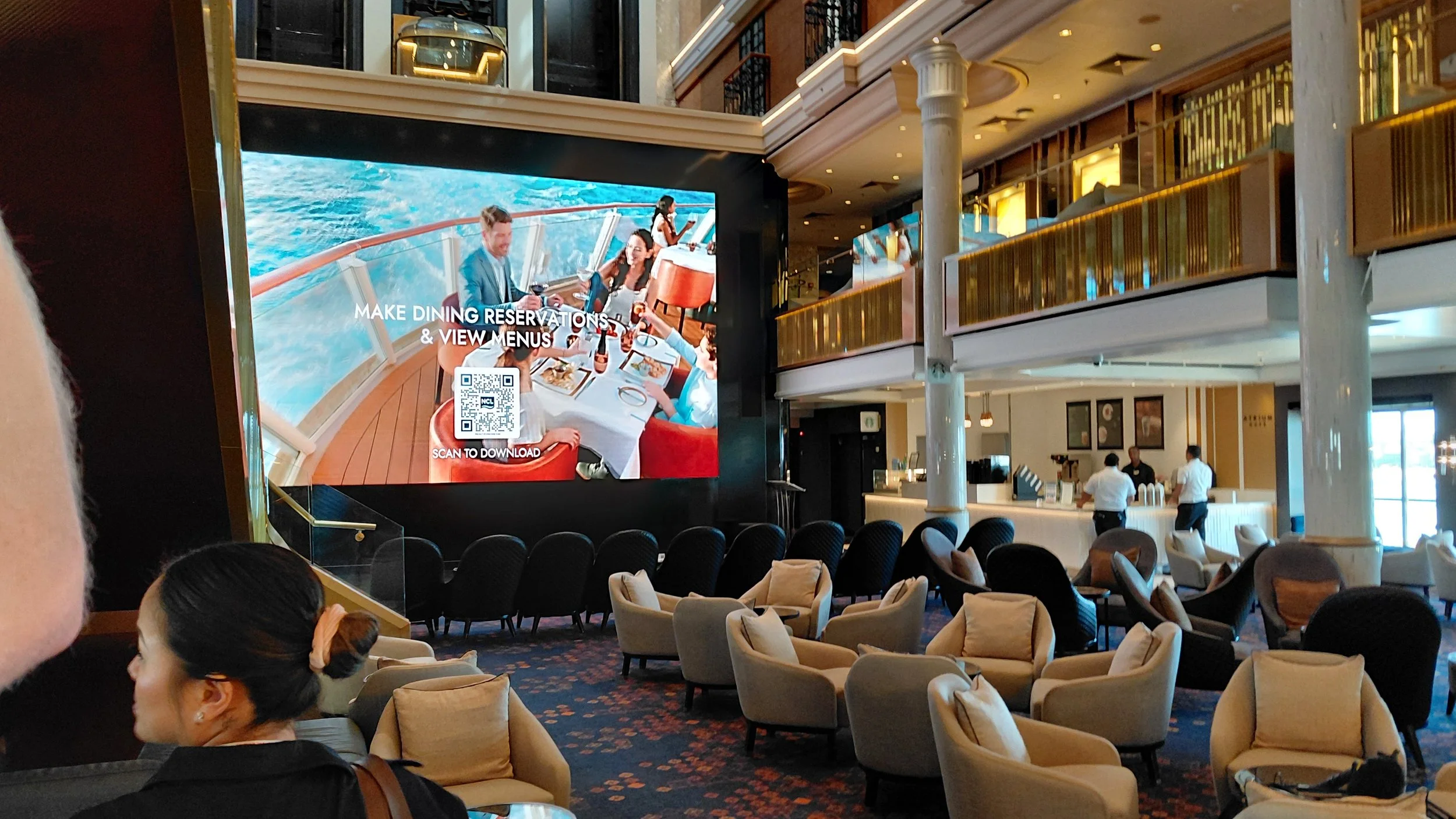 Interior of a cruise ship lobby with a large digital screen showing people dining on a ship deck, and seating area with beige chairs and some onlookers at the bar counter.