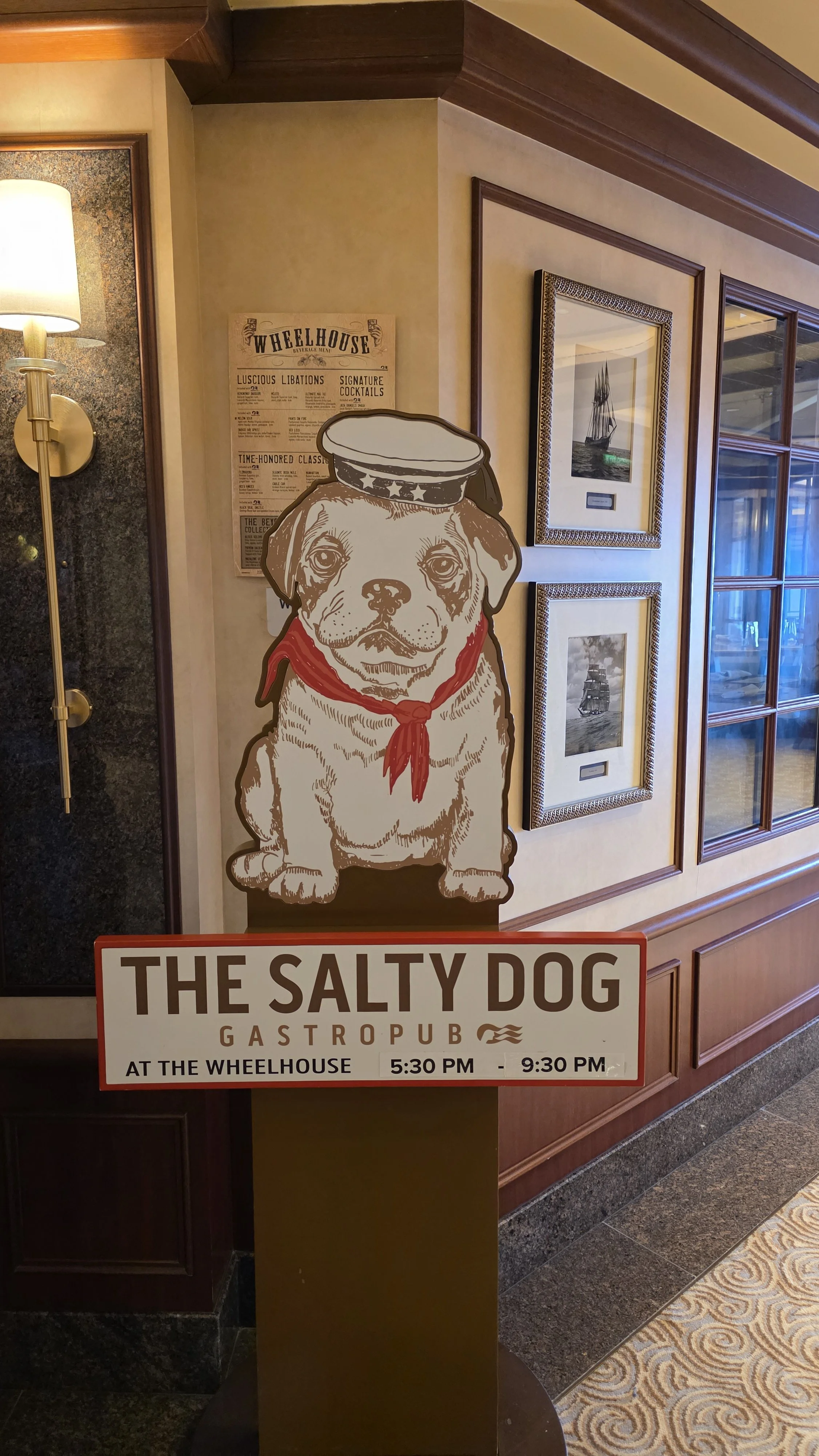 Sign with an illustration of a dog wearing a sailor hat and red bandana, advertising 'The Salty Dog Gastropub' at The Wheelhouse from 5:30 PM to 9:30 PM, in a restaurant setting with framed pictures on the wall.