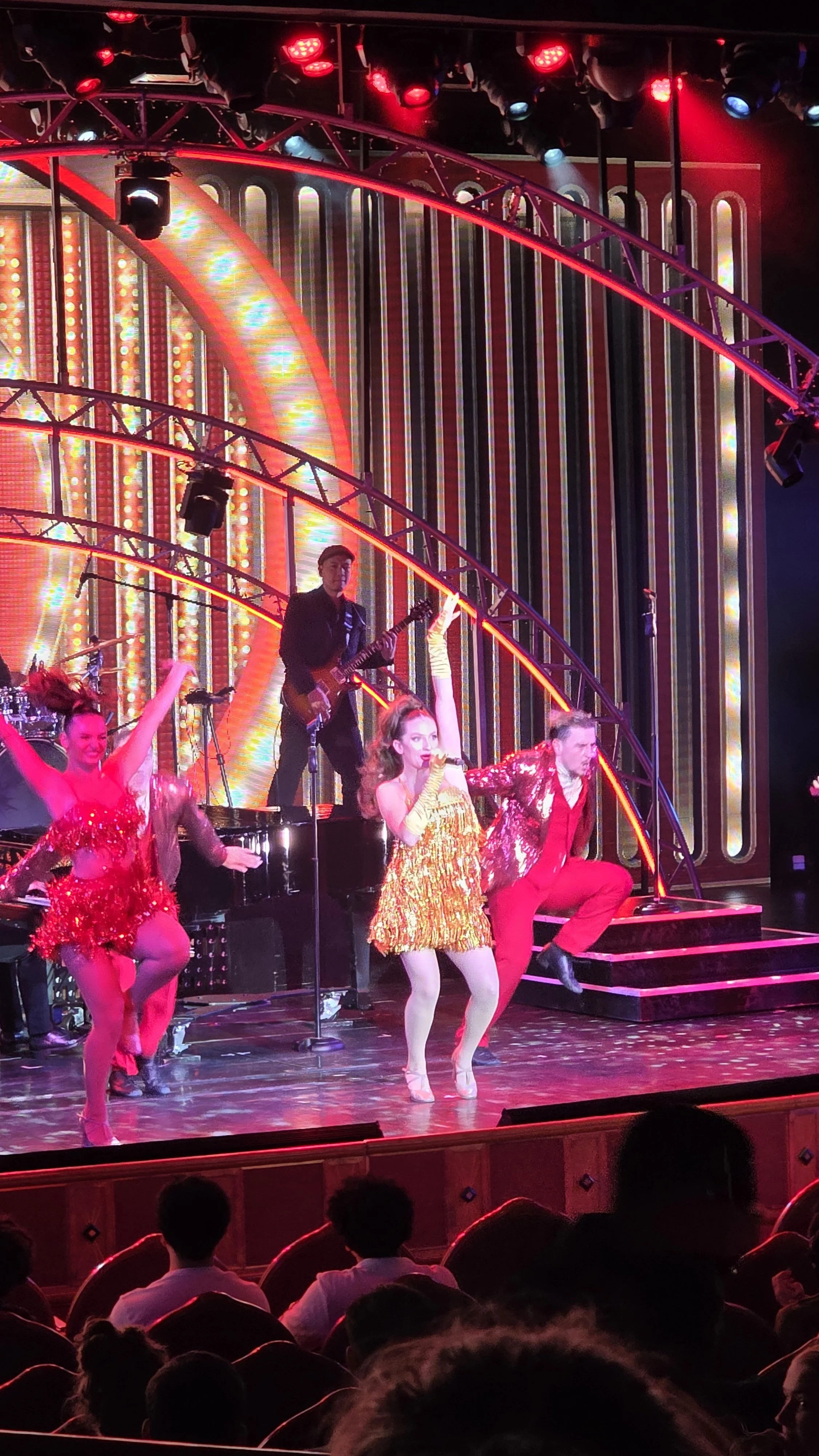 A lively stage performance with dancers and musicians dressed in vibrant, shiny costumes, performing under colorful stage lighting with a decorated backdrop.