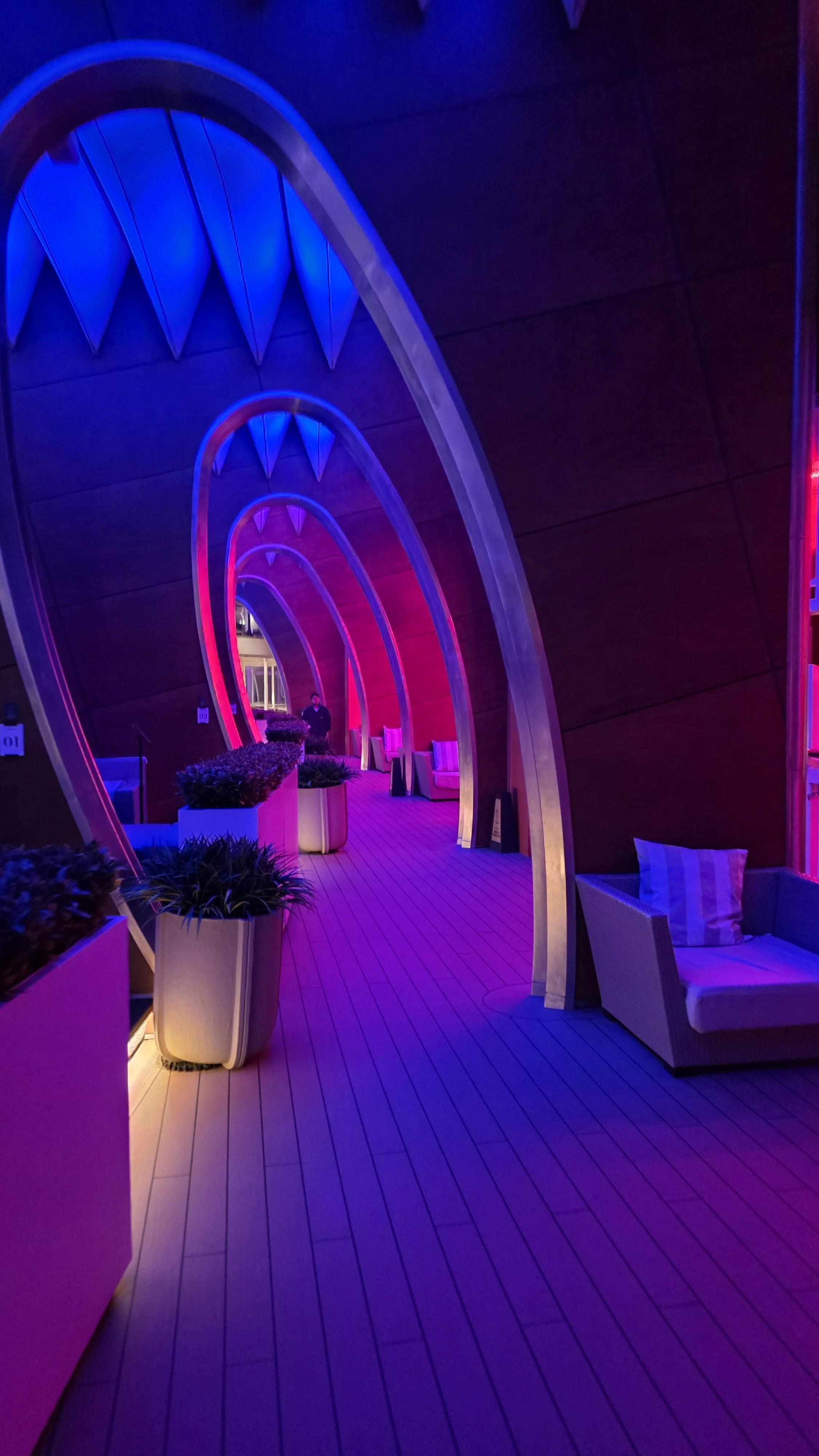Indoor hallway with arched ceilings illuminated with purple and pink lights, lined with potted plants and seating areas.