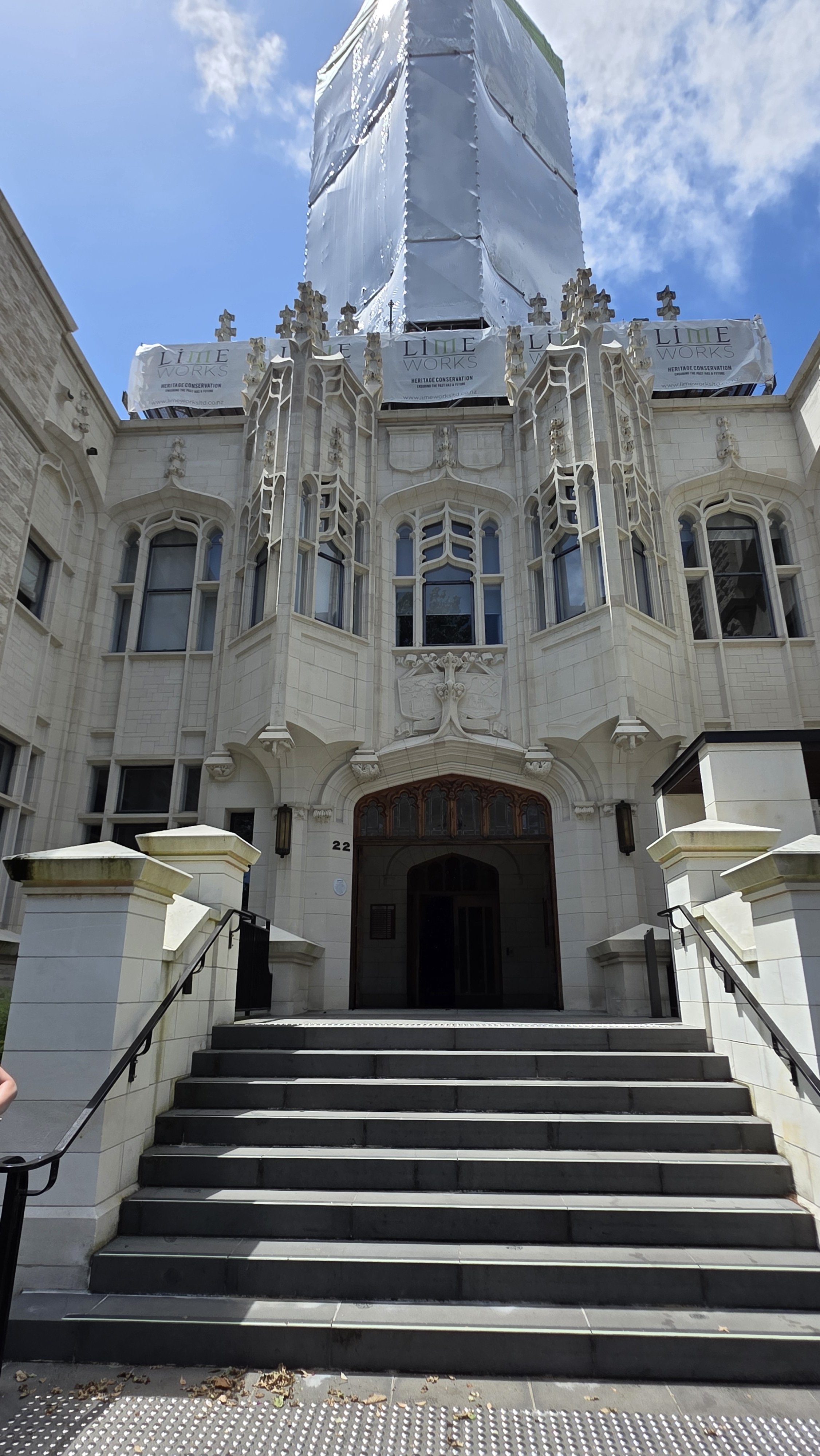 A historic building under renovation with scaffolding and protective covering at the top, featuring Gothic architectural details and a dark wooden door at the entrance.