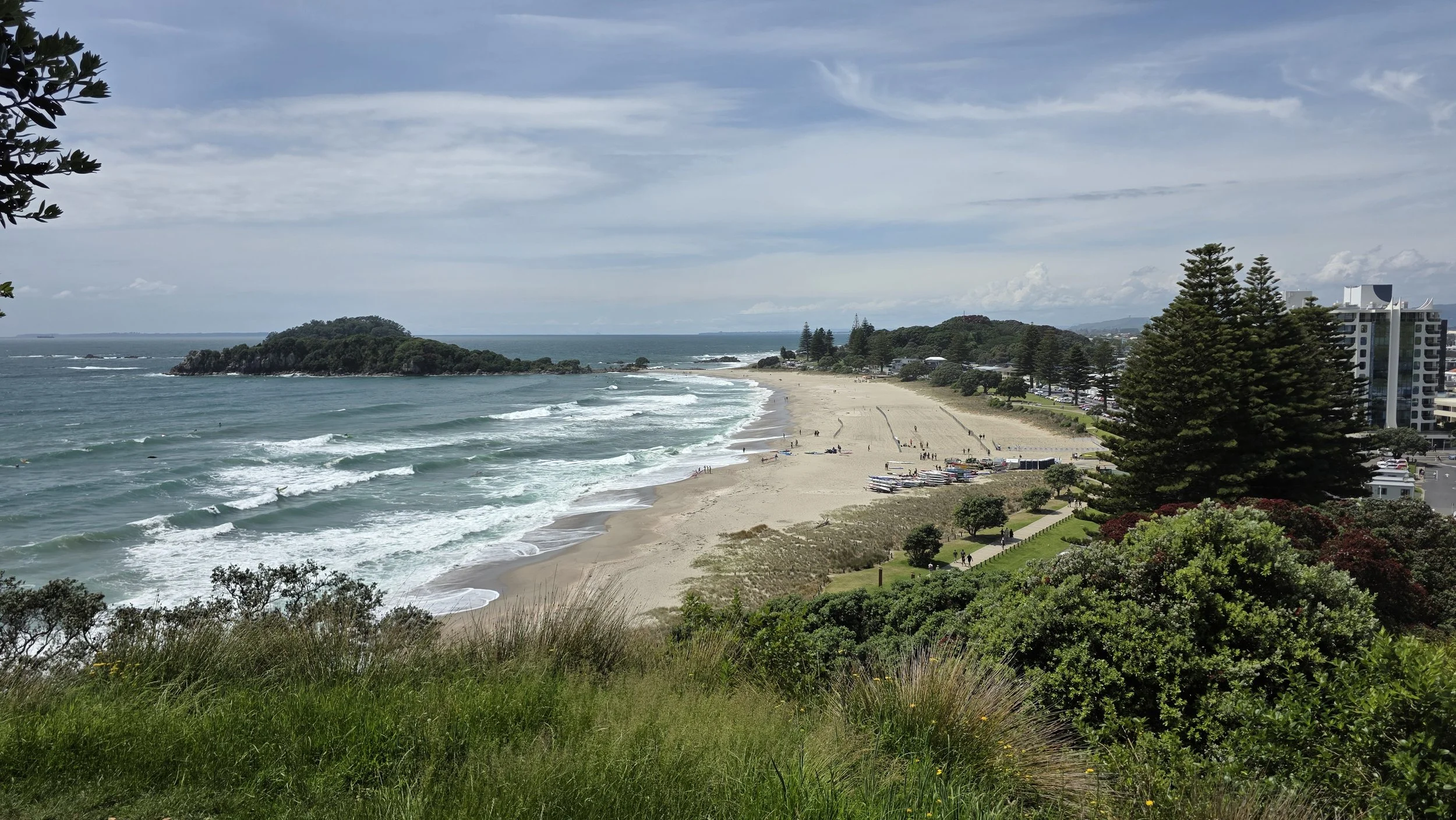 A panoramic view of a beach with a sandy shoreline, ocean waves, green vegetation and trees, and some buildings in the distance on a cloudy day.
