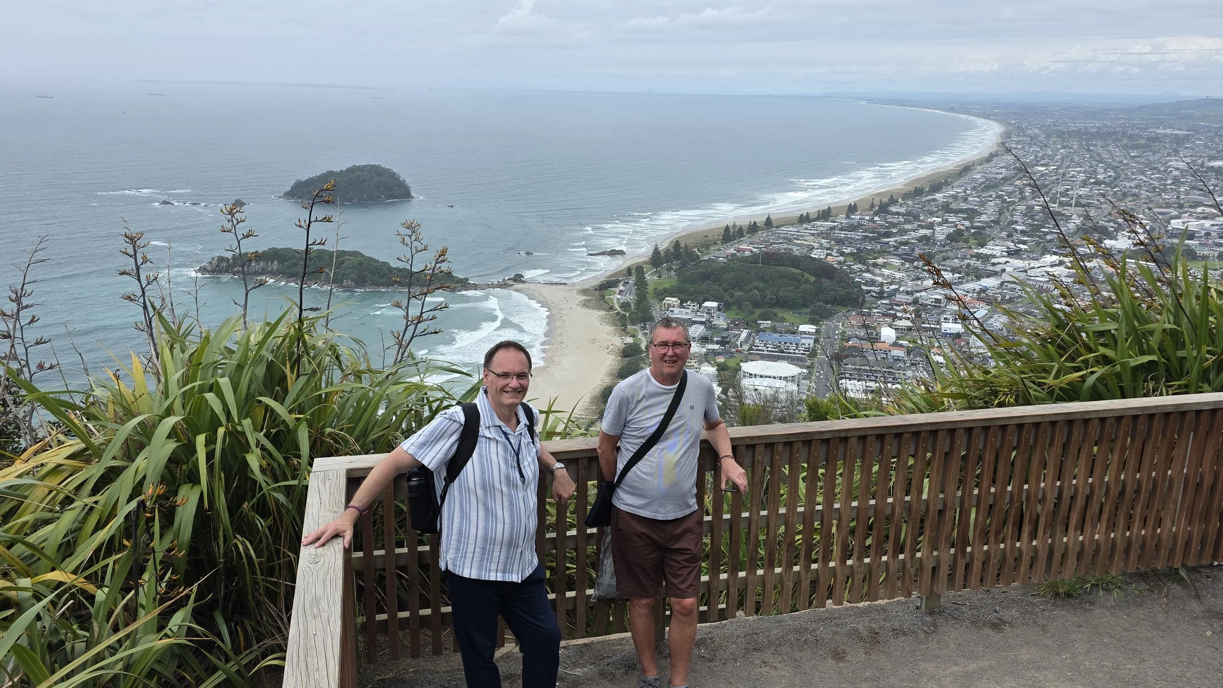 Two men smiling and posing at a scenic overlook with a view of a beach, ocean, and coastal town in the background.