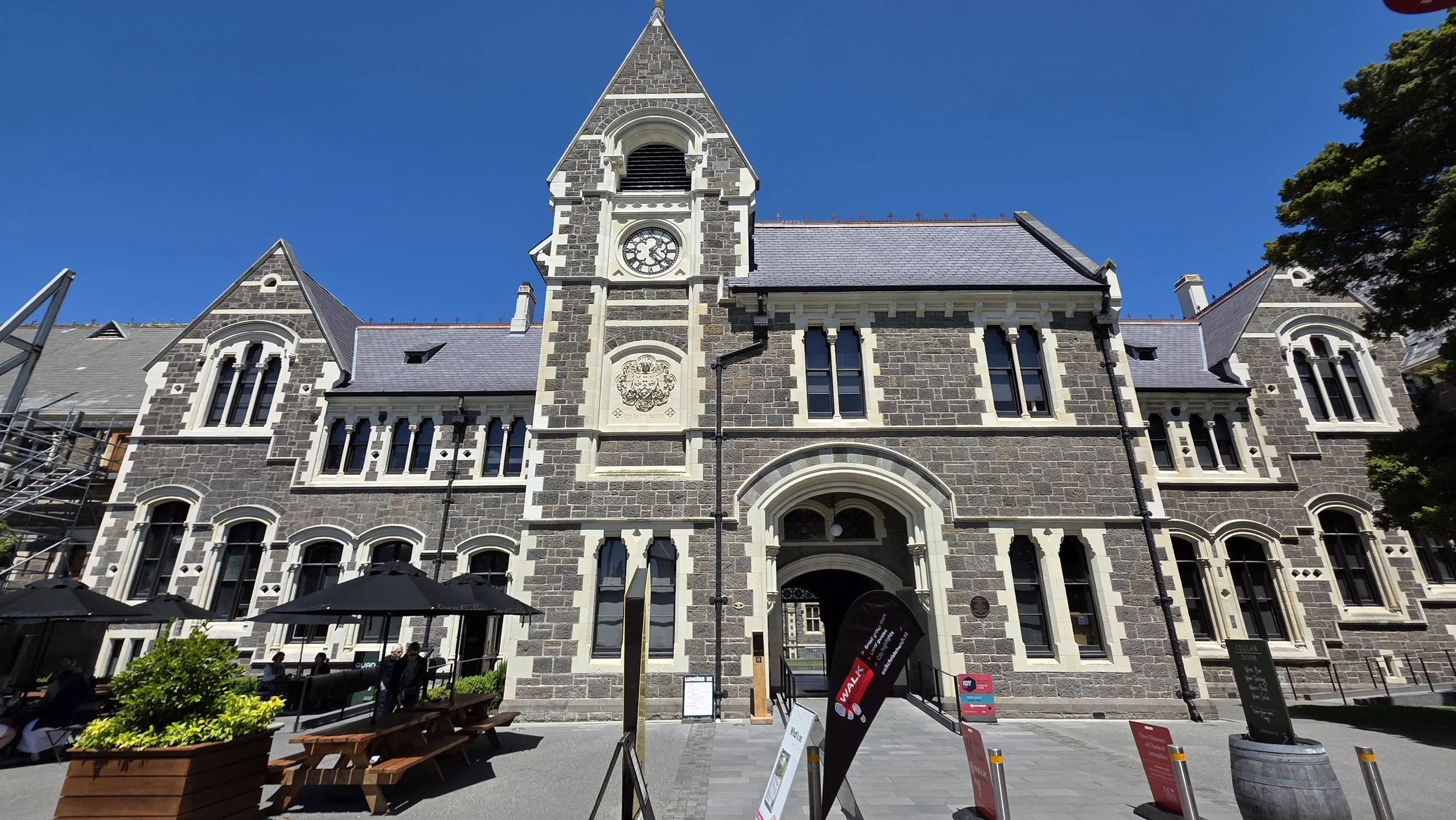 A historic stone building with a clock tower, arched windows, and a main entrance, with outdoor seating and umbrellas in front