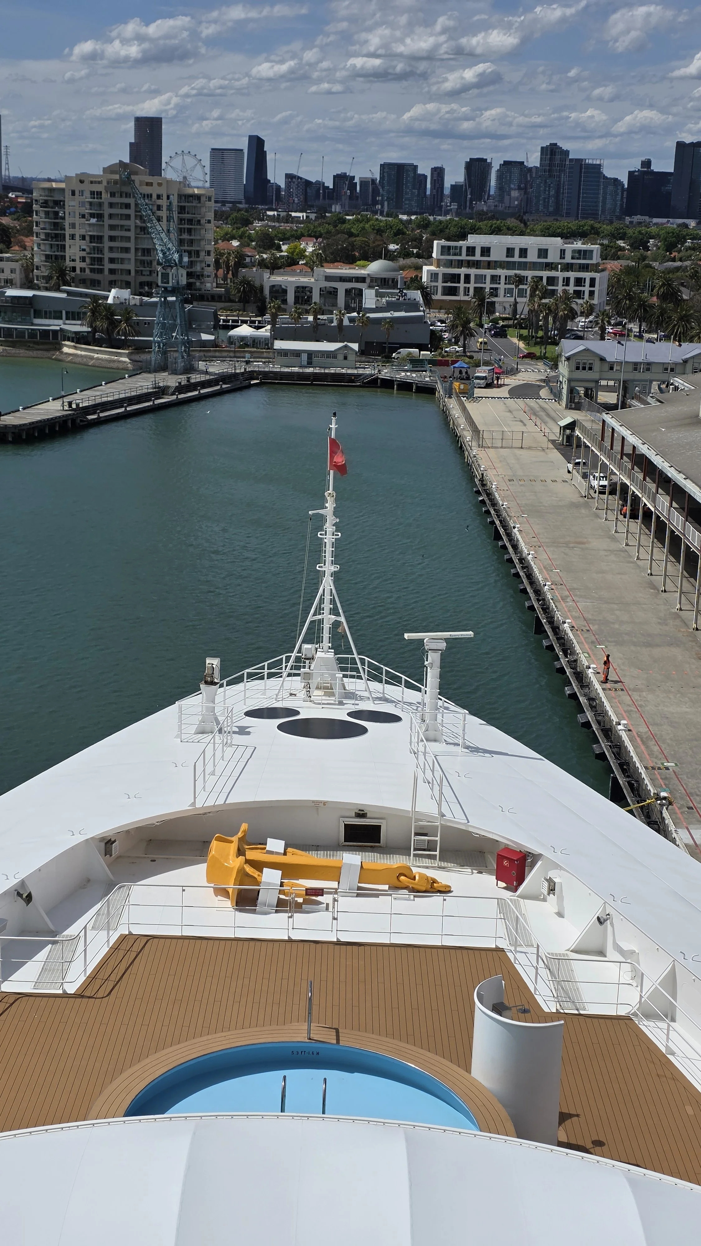 View from a cruise ship's deck showing a small pool, a yellow life raft, and a city skyline with tall buildings in the background.