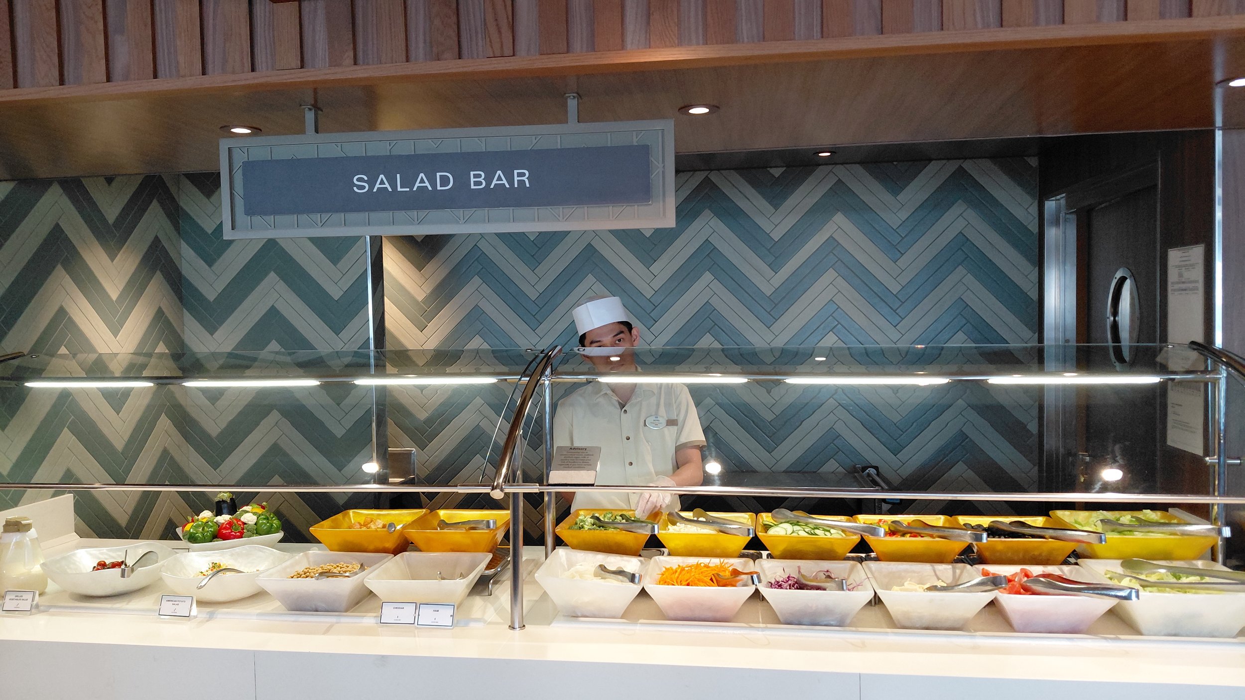 A salad bar with various bowls of fresh vegetables, and a worker in a white uniform and hat behind the counter.