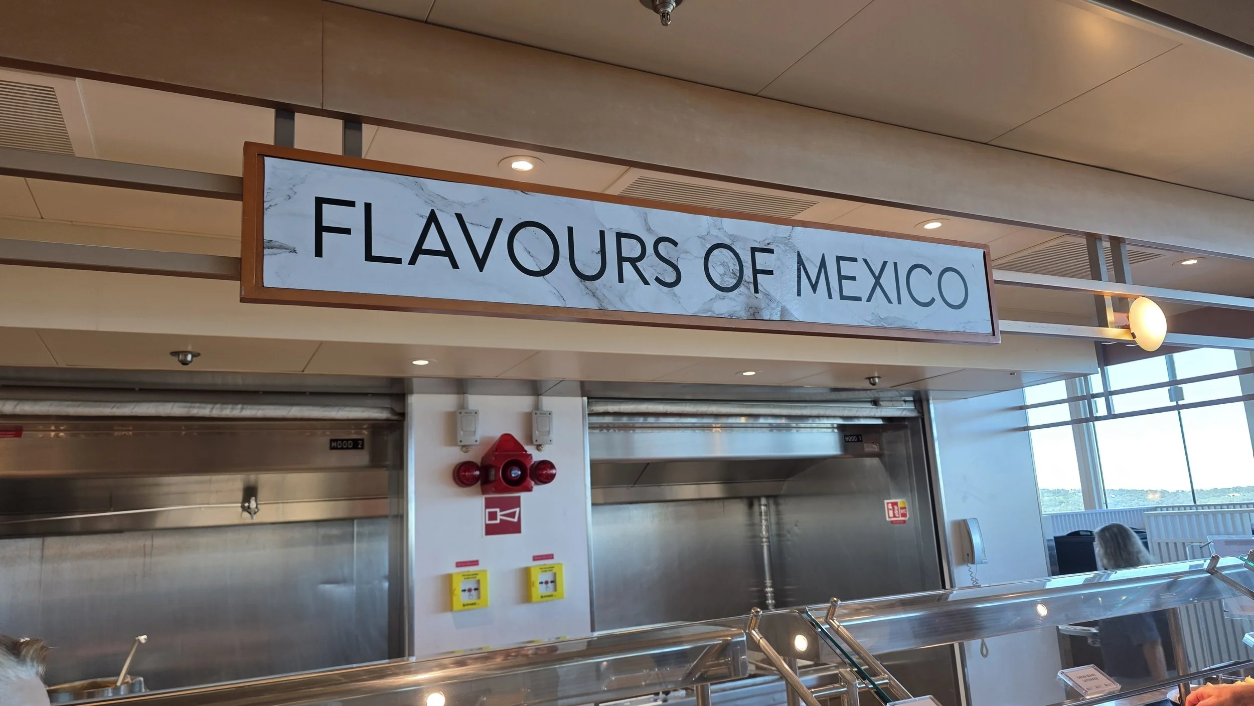 Sign reading 'Flavours of Mexico' at a food counter or restaurant with stainless steel surfaces and a view of windows in the background.