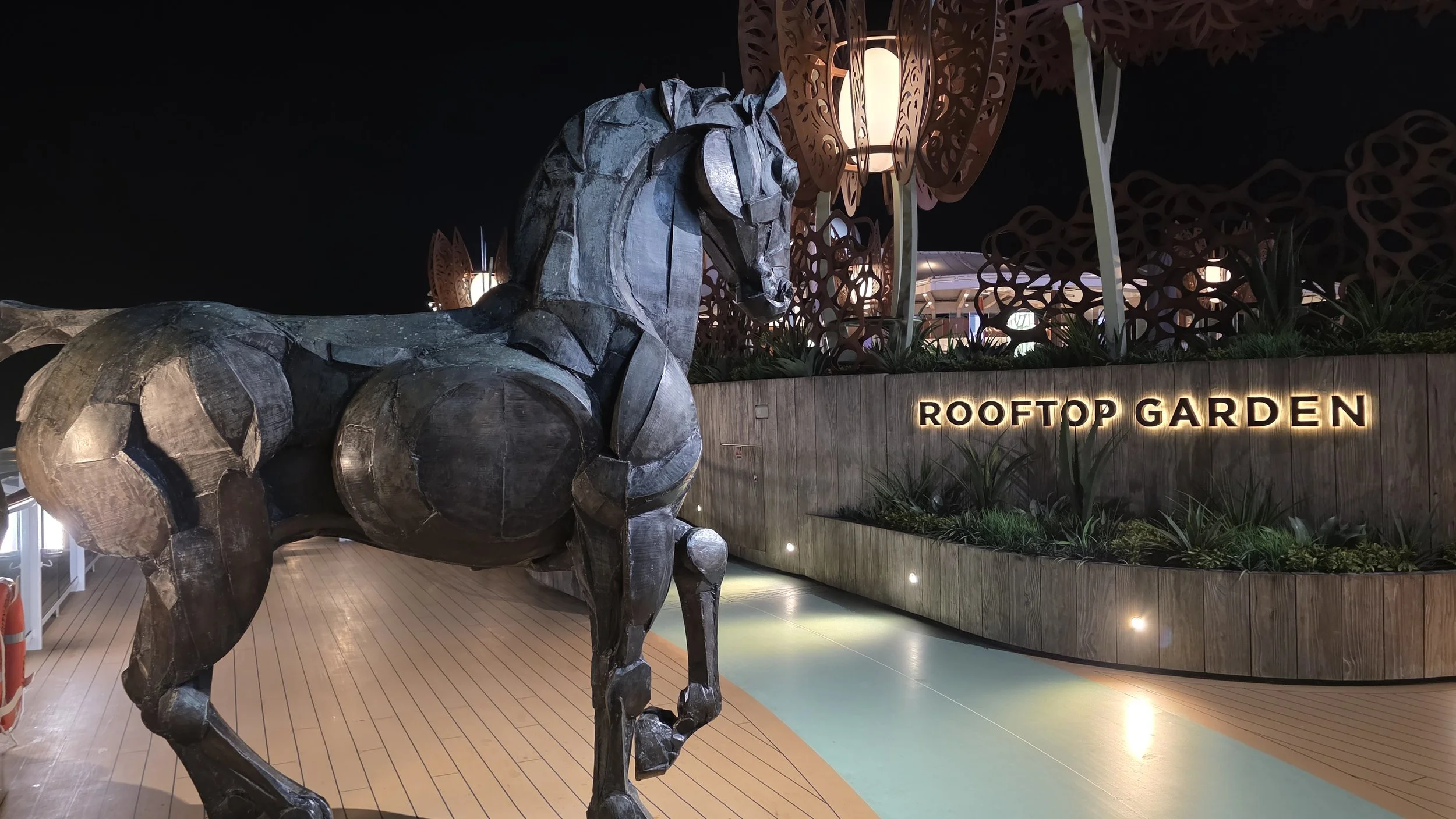 A large, dark-colored wooden sculpture of a horse standing on a rooftop garden at night, with illuminated signs reading 'ROOFTOP GARDEN' in the background.