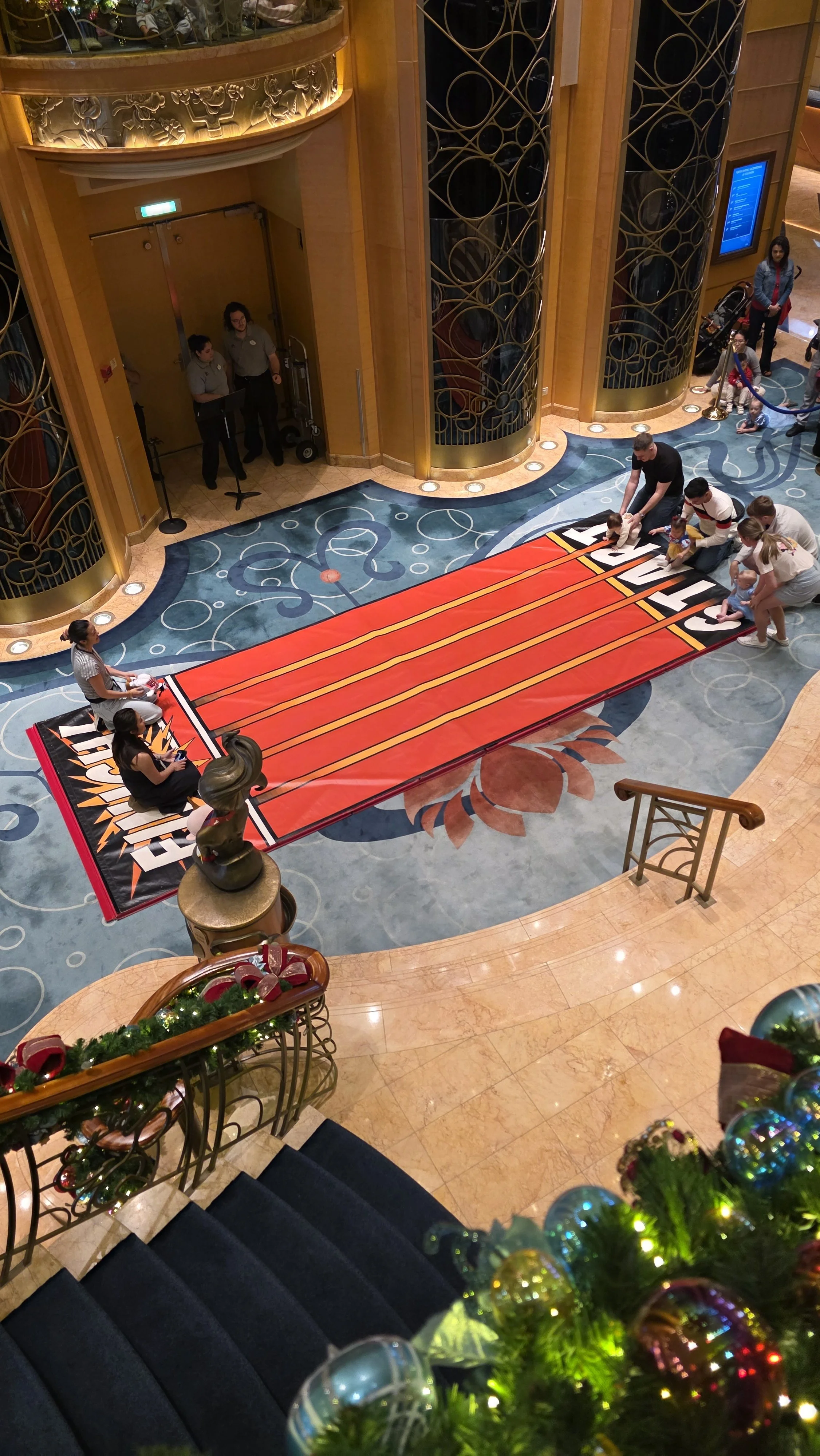 An indoor scene with a large red and orange competition-style treadmill on a decorative carpet. Several children and adults are engaging with the treadmill, with some children lying on it and adults supervising. The background features elegant gold a
