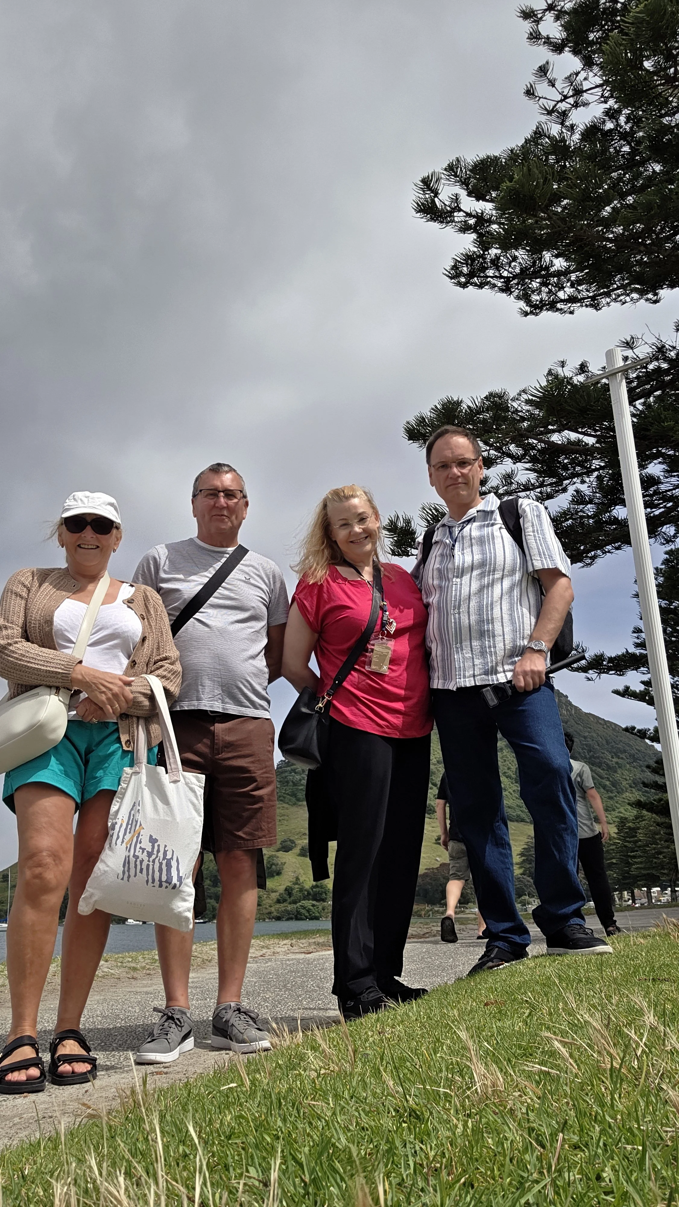 Four people standing outdoors on a cloudy day near a body of water with a mountain in the background. They are smiling and dressed casually, with some carrying bags and wearing sunglasses.