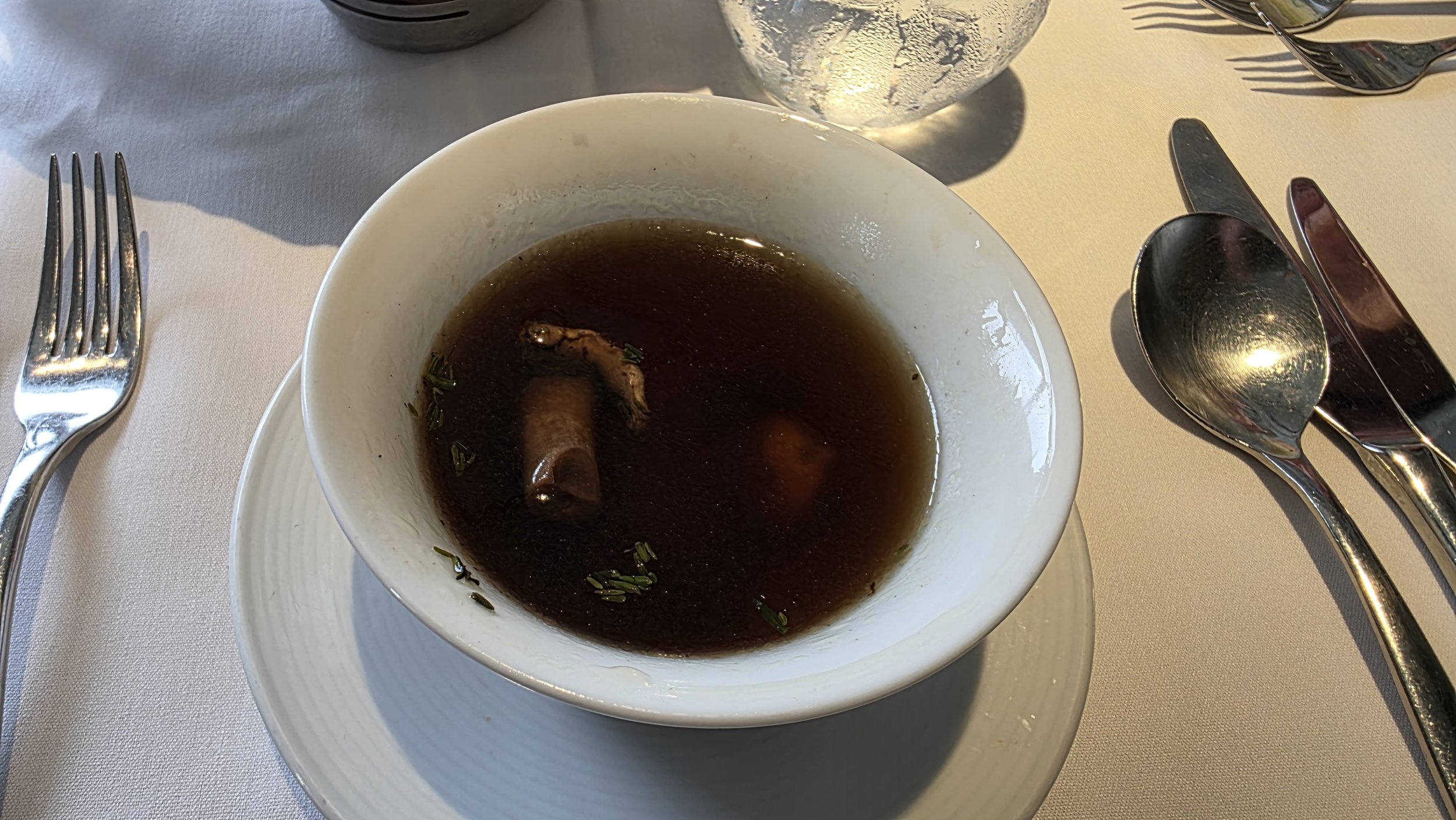 Empty bowl of dark broth with a mushroom and small herbs on a dining table, with silverware and a glass of water.