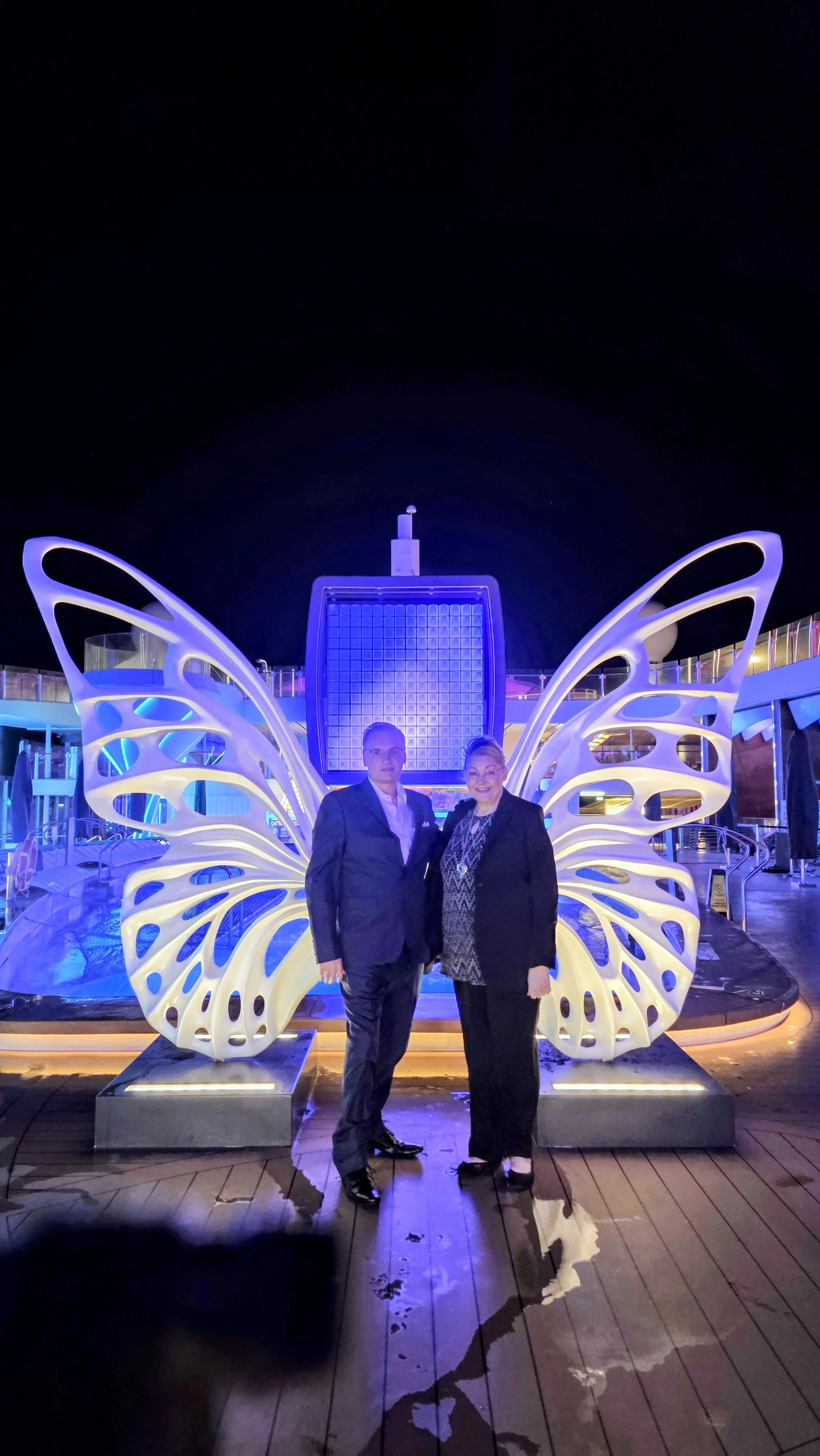 Man and woman in formal attire standing together on a deck with illuminated butterfly wings and a large illuminated square sculpture with a small tower on top in the background, against a dark night sky.