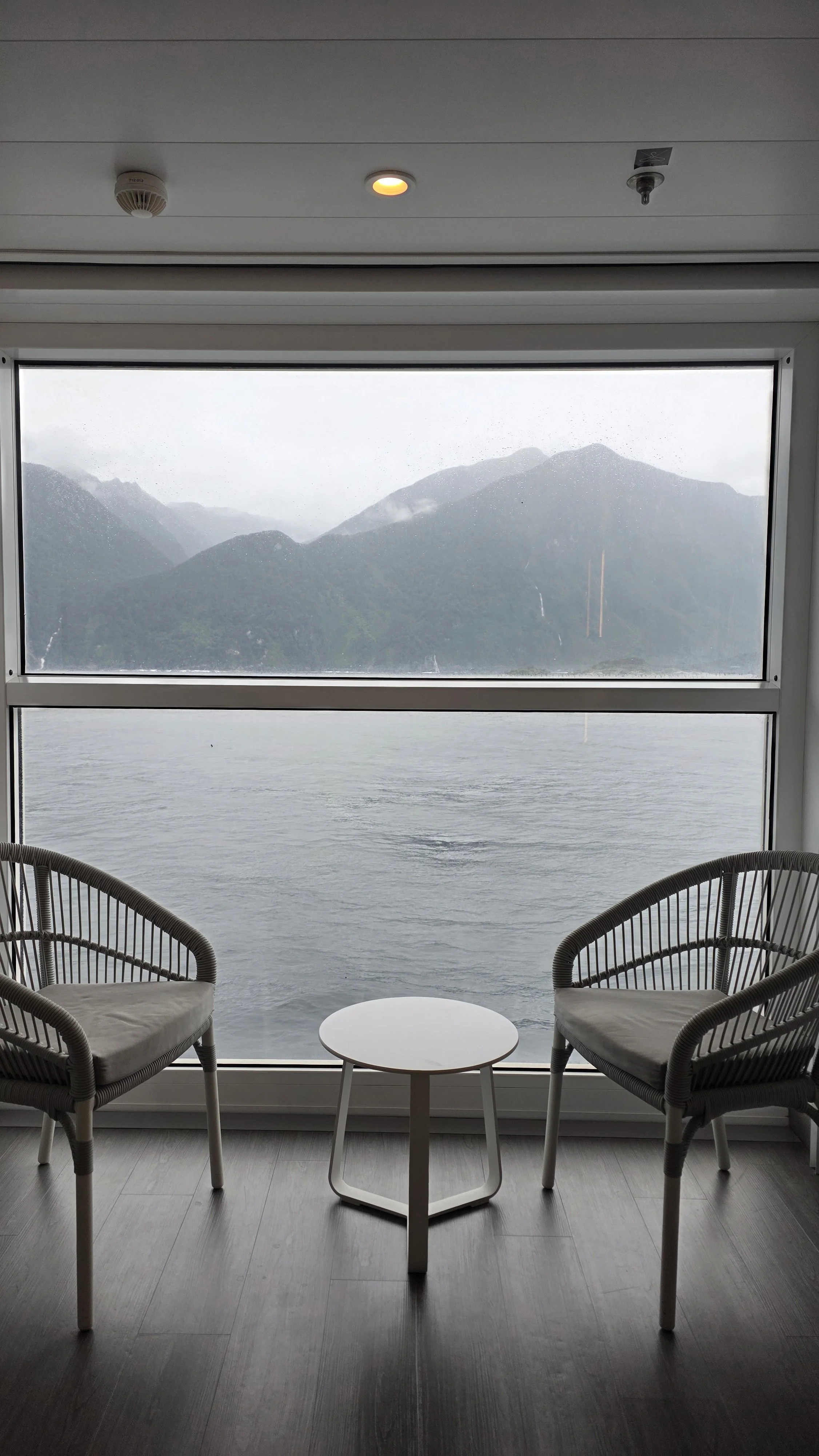 A cozy indoor space with a large window view of a gray lake and mountains, two modern chairs with light cushions facing the window, and a small white round table between them, on a gray wooden floor.