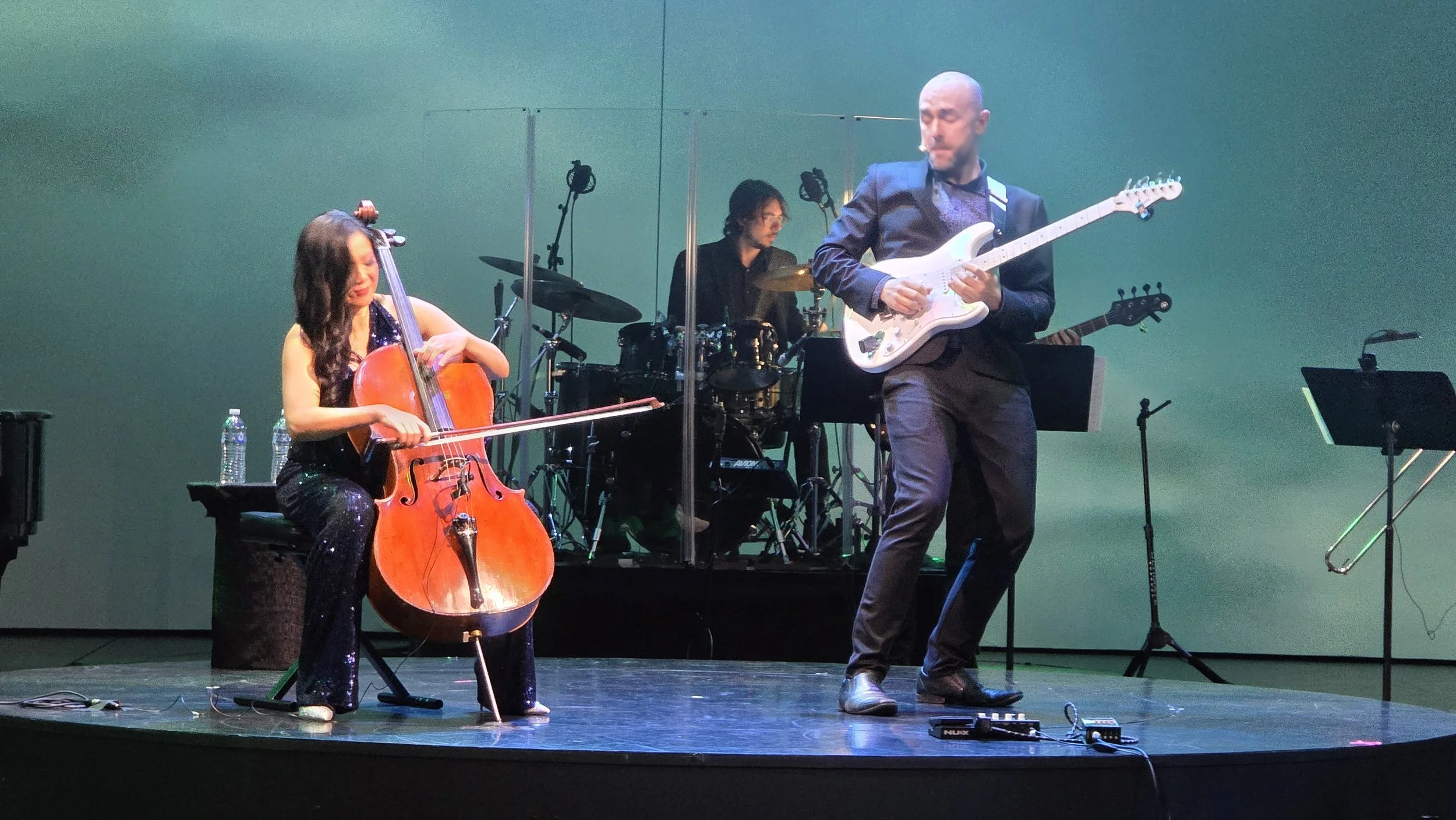 Musicians performing on stage with a woman playing a cello, a man playing an electric guitar, and a drummer in the background, all behind a transparent screen.