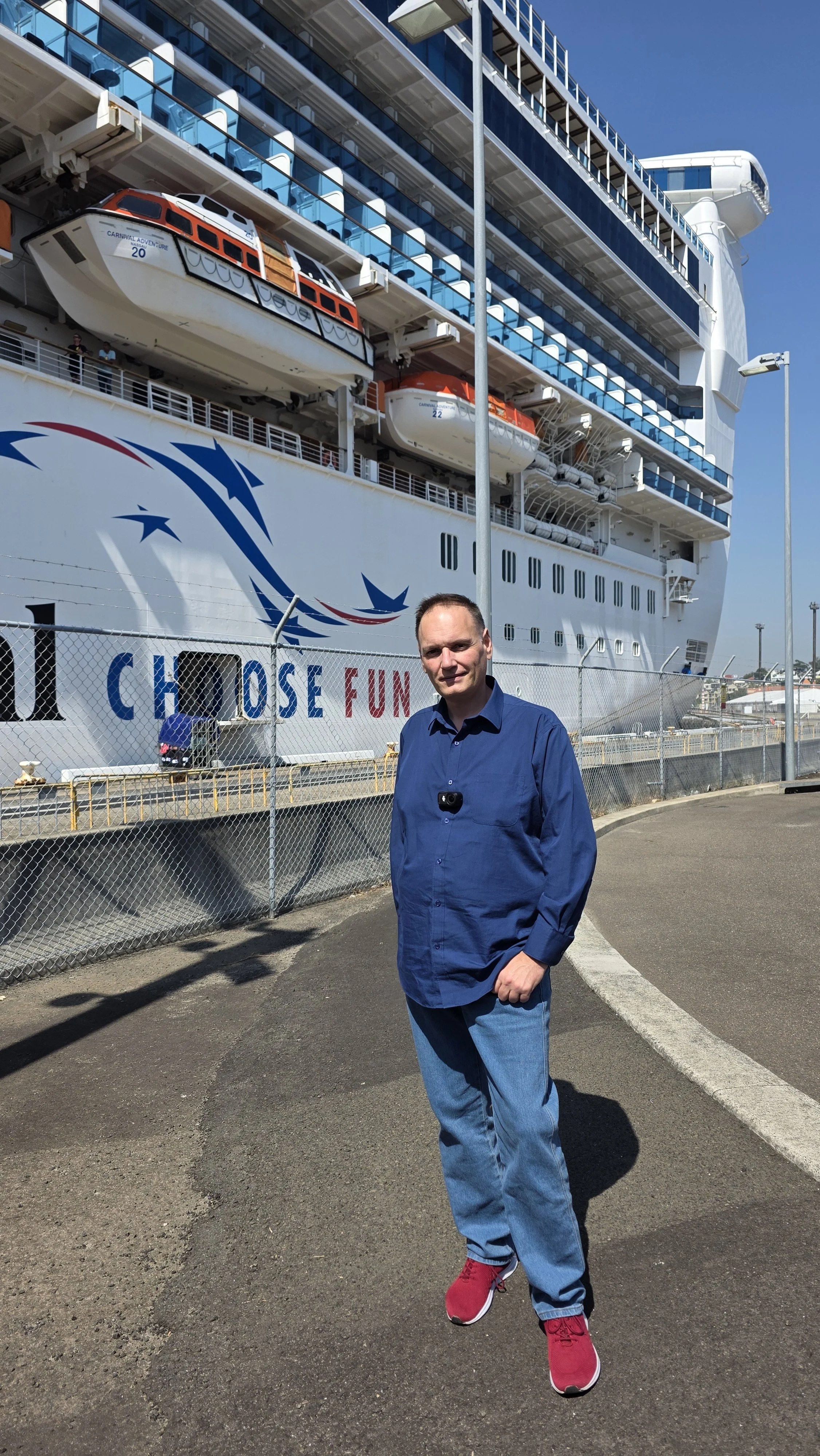 A man in a blue shirt and jeans standing in front of a docked cruise ship with lifeboats visible on the side, and the ship's name partially visible with the words 'Choose Fun'.