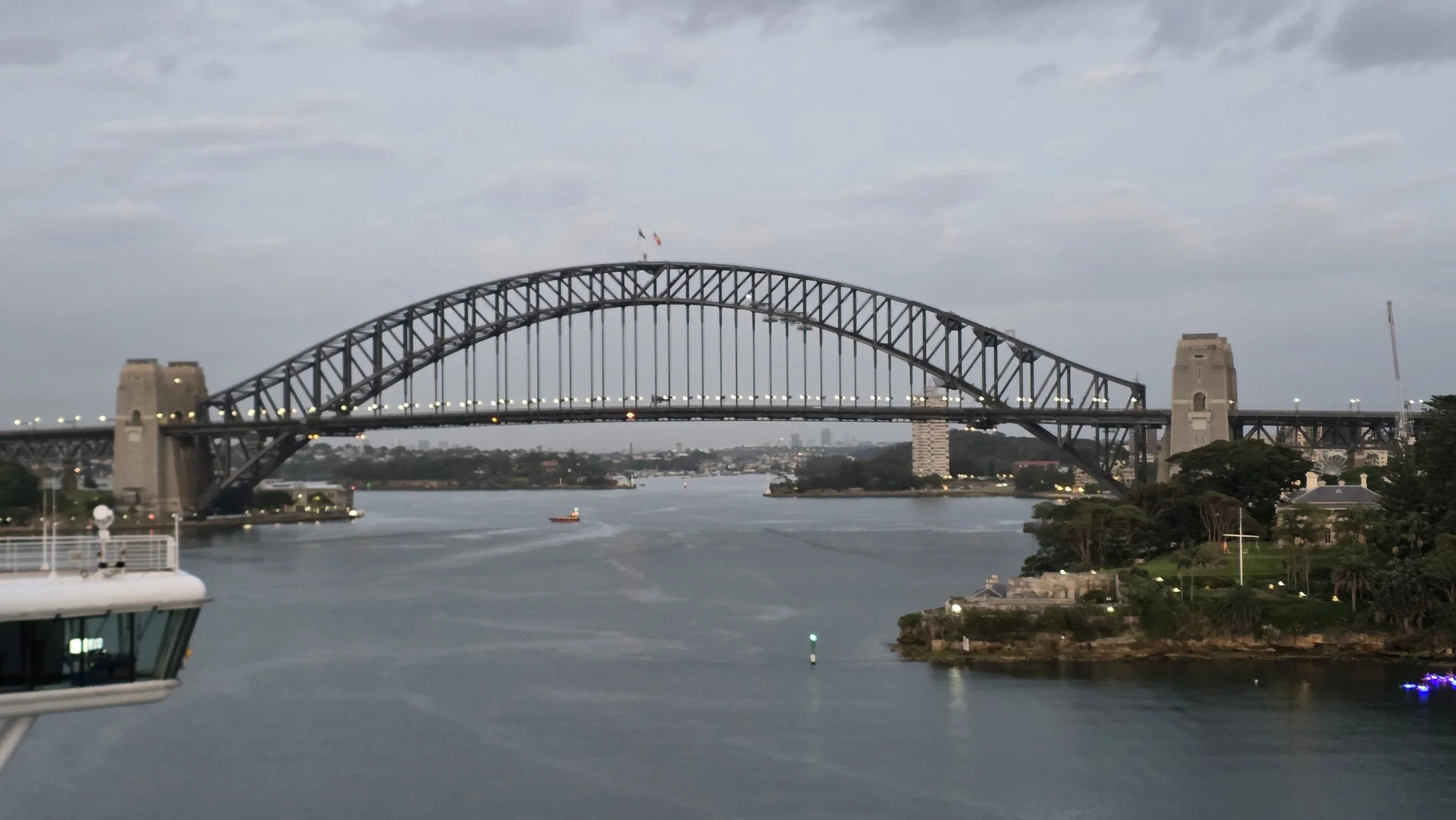 Sydney Harbour Bridge over water with a boat, city skyline in background, and cloudy sky.