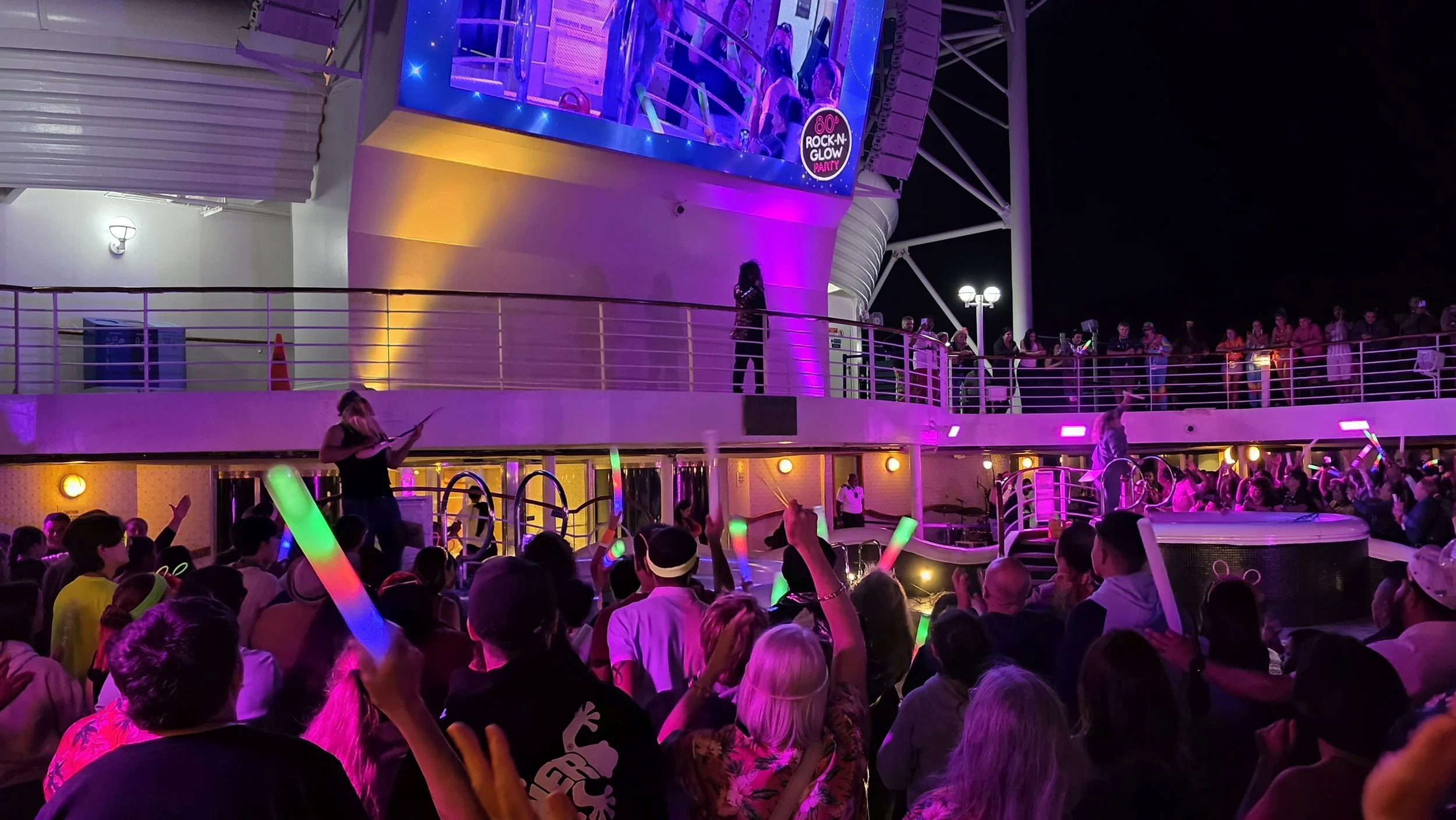 Crowd of people on a cruise ship deck at night, dancing and waving glow sticks, with colorful lights and a large screen projecting the scene.