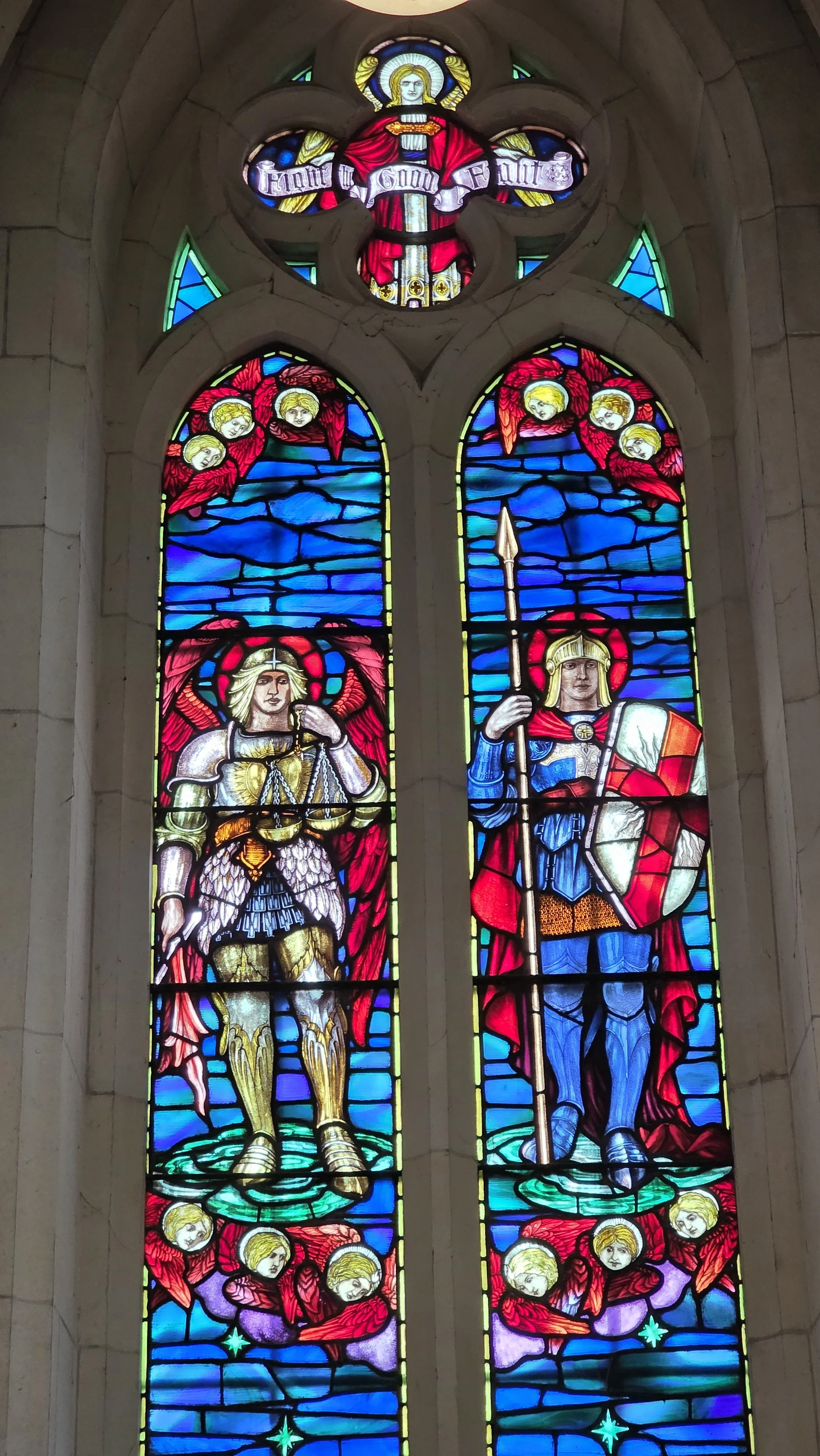 Colorful stained glass window depicting two saints, with cherubs and religious symbols and inscriptions in vibrant red, blue, and yellow colors.