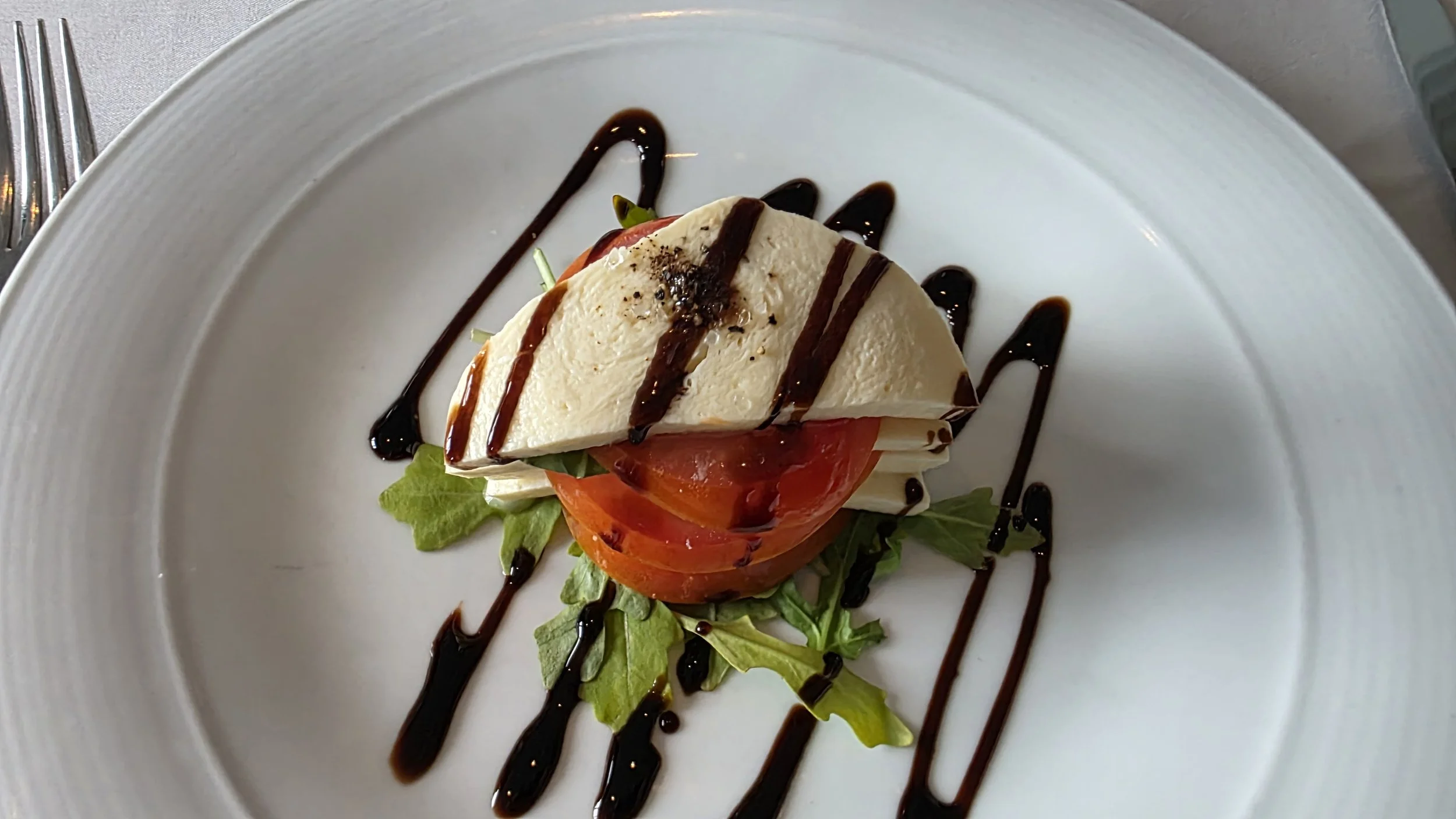 A plate with sliced tomato, fresh greens, and sliced mozzarella cheese, garnished with balsamic glaze.