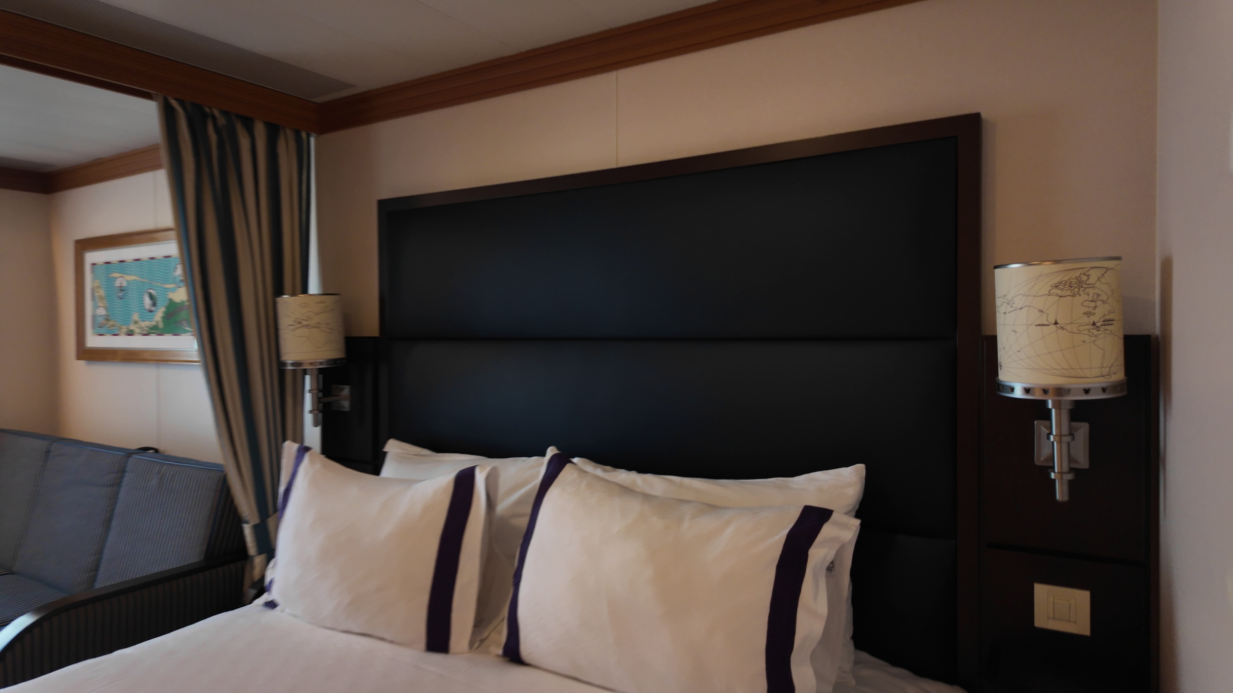 Hotel bedroom with a large bed, black headboard, and white pillows with purple borders. Wall-mounted lamps on either side of the bed, a framed map on the wall, and striped curtains partially open revealing a window.
