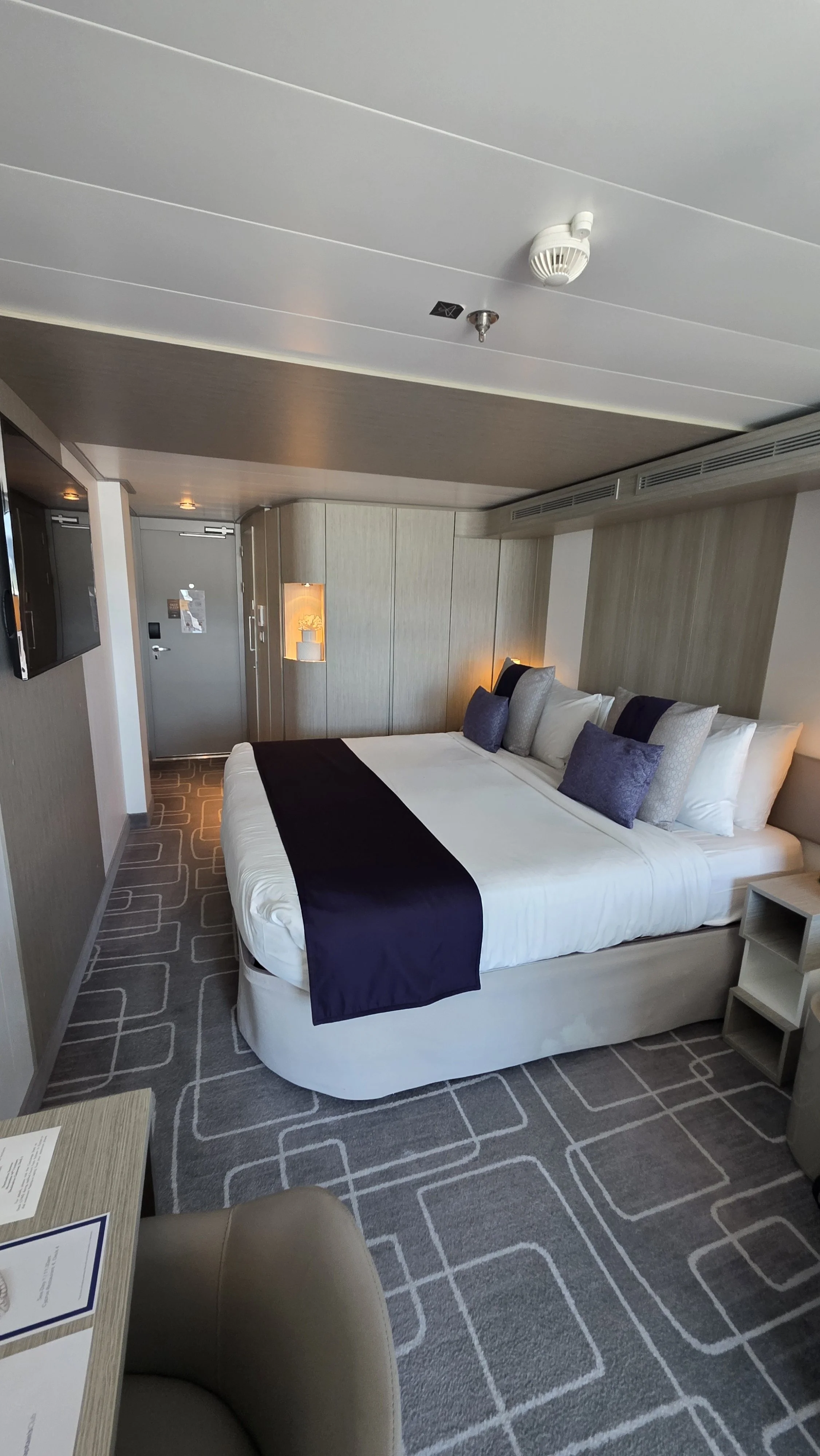 A hotel room with a large bed topped with multiple pillows, a navy blue bed runner, and modern decor. There is a wall-mounted TV, a nightstand, and a carpet with geometric patterns. The room's door and a small illuminated shelf are visible in the bac