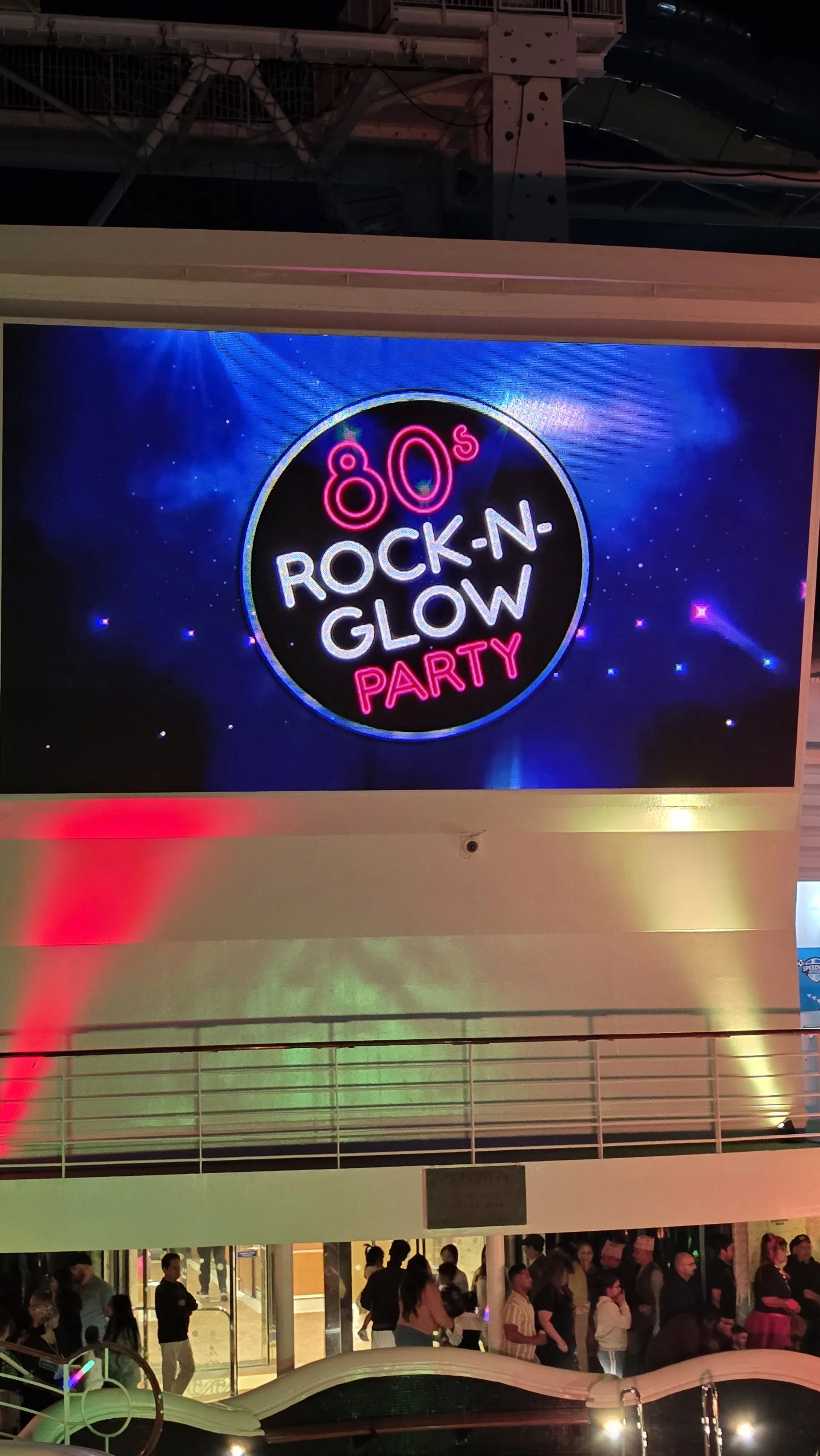 A digital screen displaying a neon sign that reads '80s Rock-N-Glow Party' with a space-like background, pulled above a crowd of people at an indoor event.