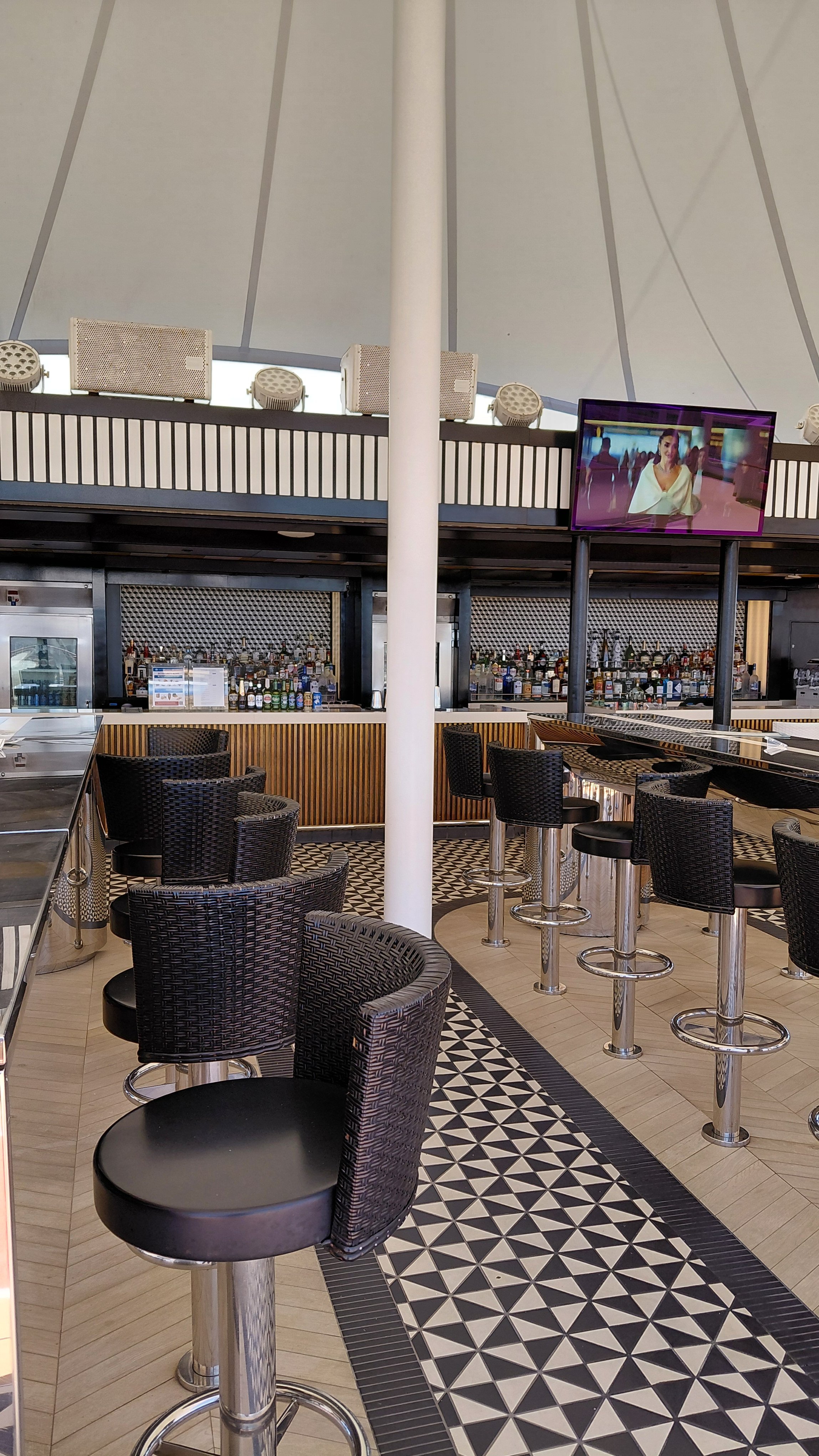 Empty bar area with black wicker chairs, high tables, and a patterned tile floor, with a TV screen displaying a woman and a bar with various bottles of alcohol in the background.