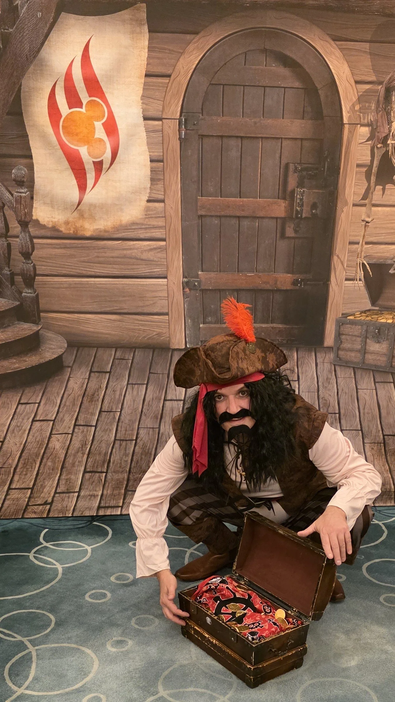 Person dressed as a pirate with a black beard and curly hair, wearing a brown hat with a red feather, squatting next to a treasure chest on a patterned blue carpet. The background features a painted wooden door and a flag with red flames and an orang
