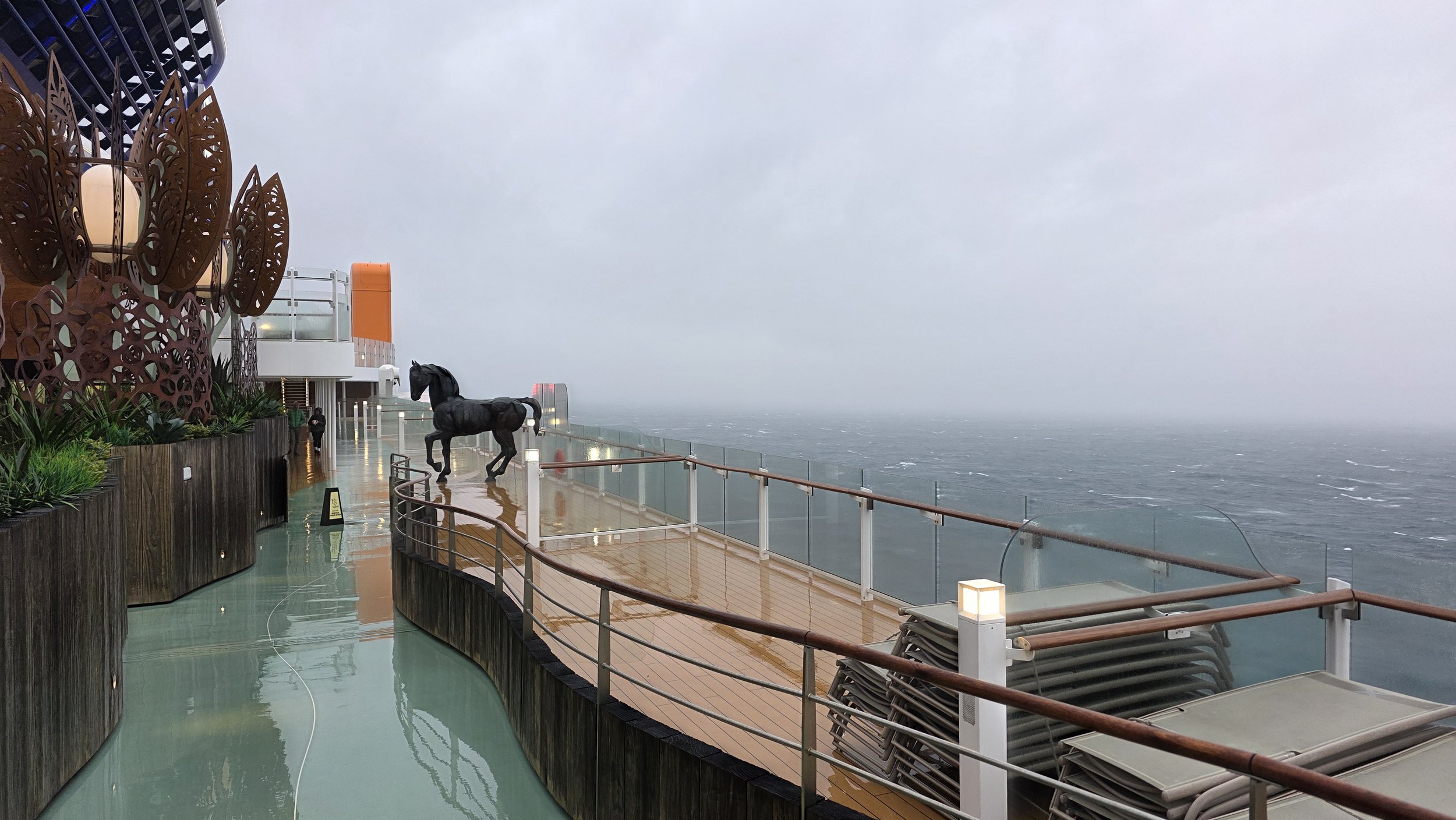 Rainy deck of a cruise ship with a black horse sculpture, stack of lounge chairs, and a view of the ocean in the background.