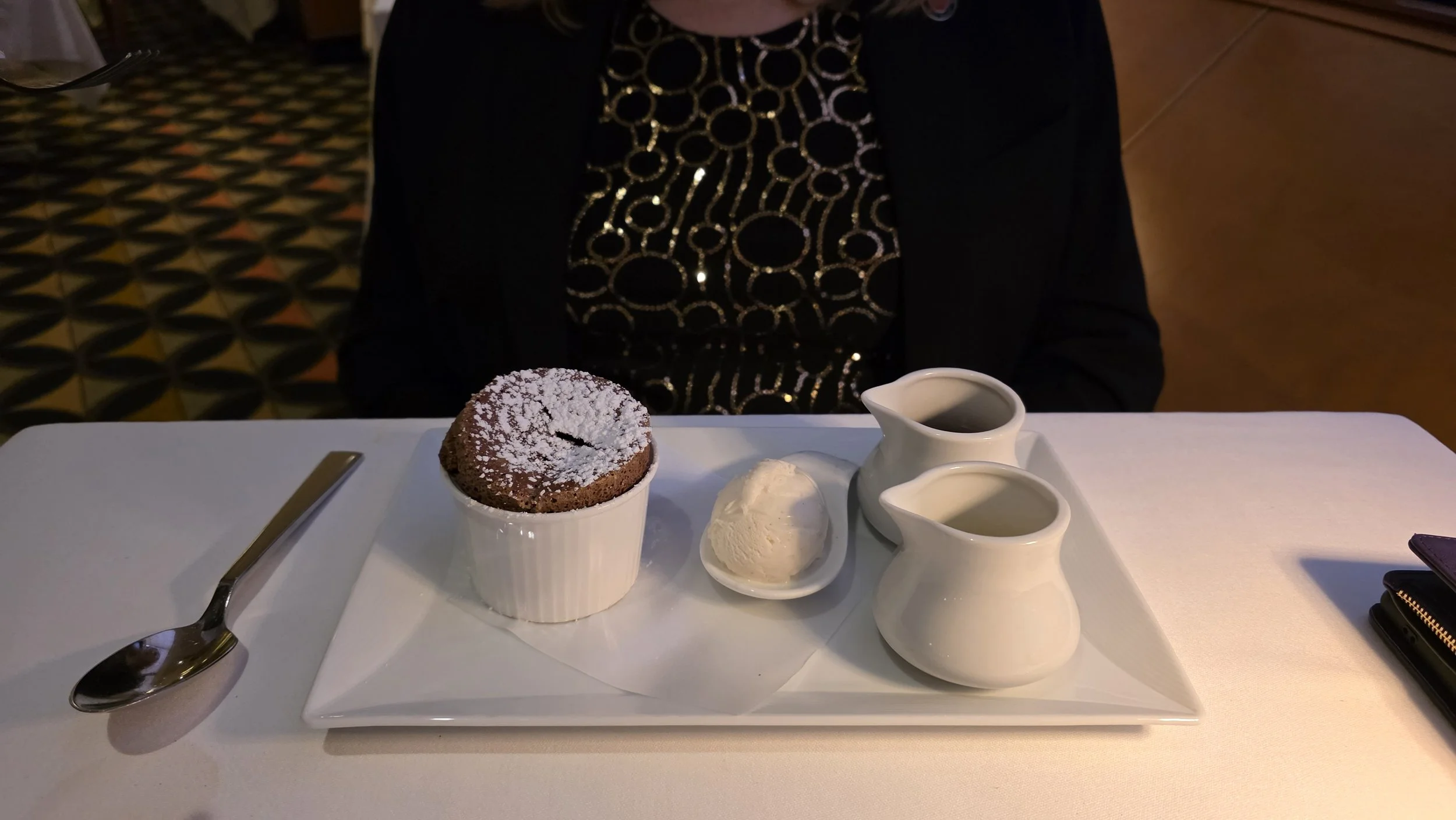 Chocolate souffle with powdered sugar, vanilla ice cream, and two small pitchers of cream or sauce on a white tray on a white table in a restaurant.