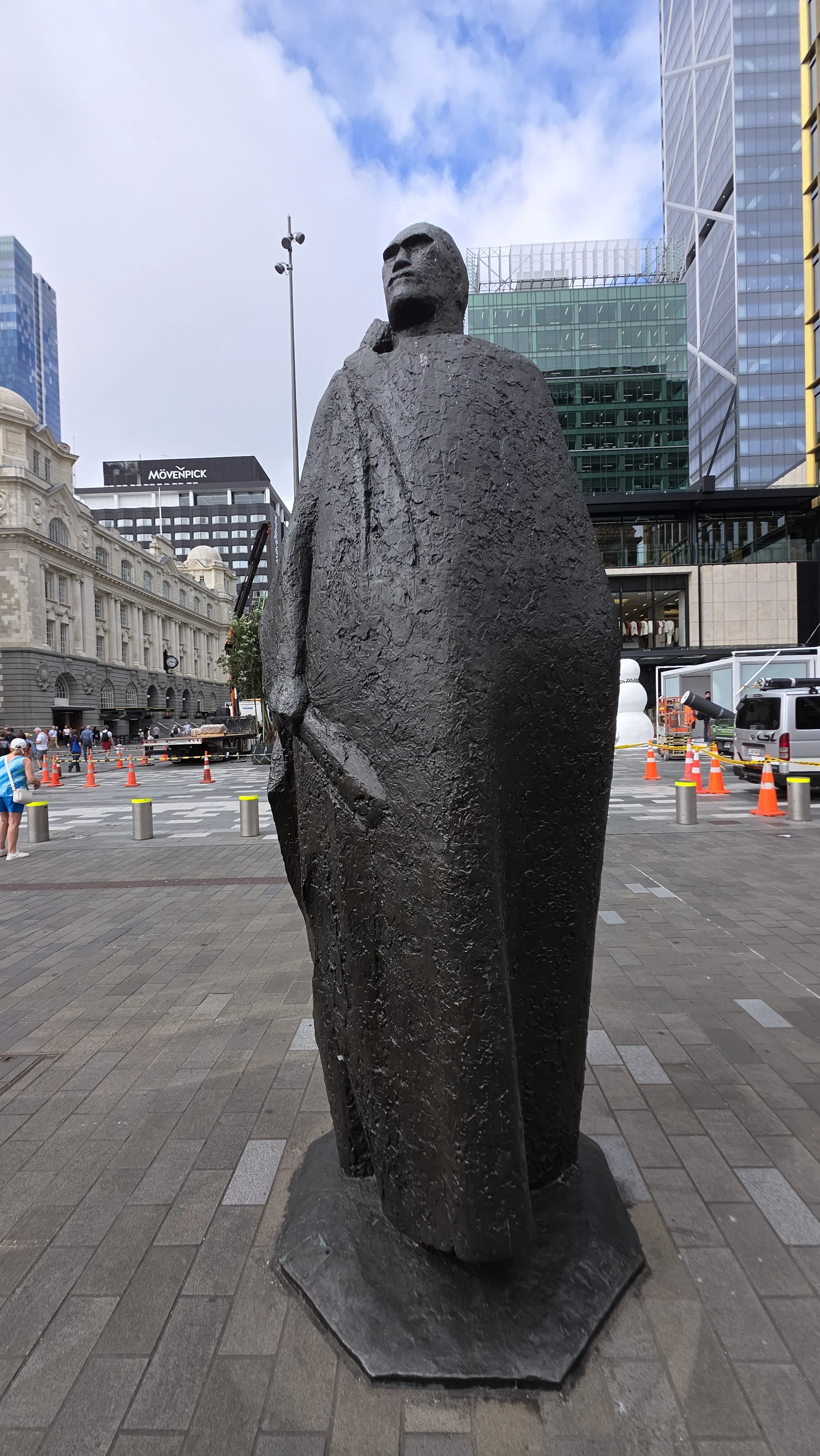 Bronze statue of a tall person wearing a long cloak, holding an object in their right hand, in an urban plaza with modern buildings and a few people visible in the background.