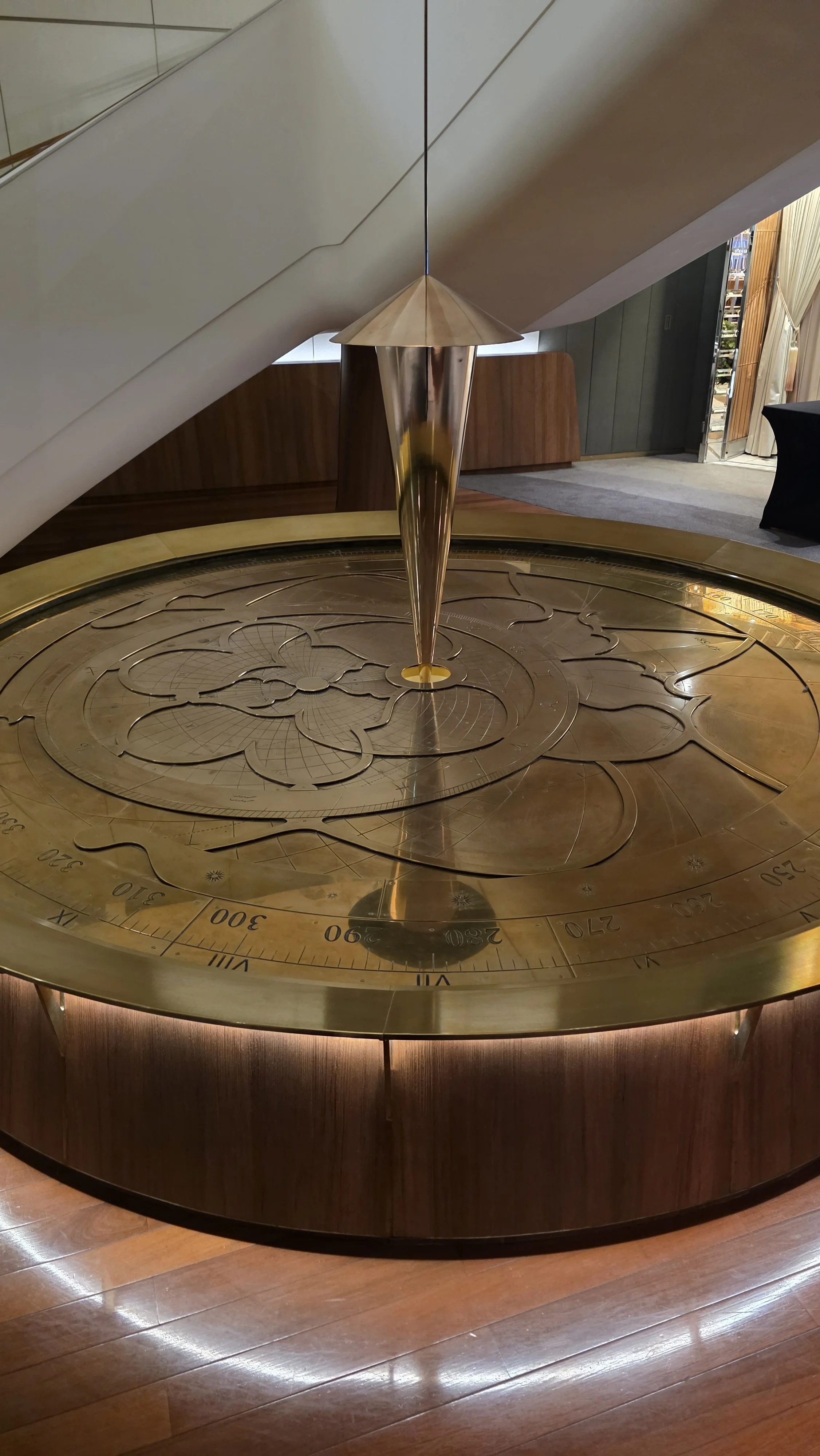 A decorative brass sundial with engraved markings and floral patterns, located indoors near a staircase with a curved wall and wooden flooring.