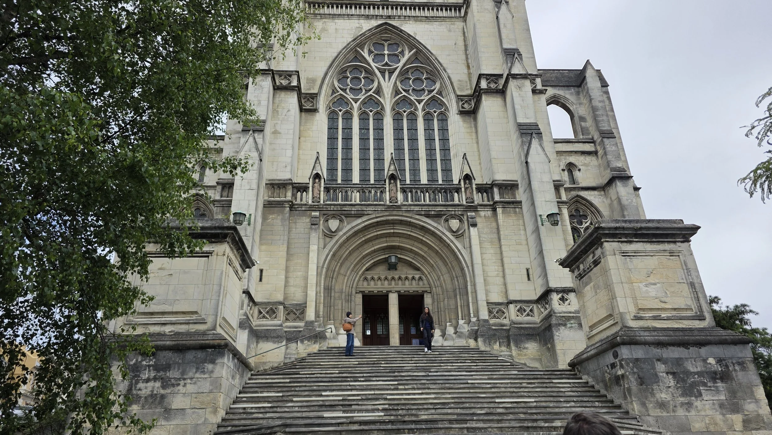 Front view of a historic stone church with gothic architecture, featuring large stained glass windows, intricate stone carvings around the entrance, and several steps leading up to the doorway. Two people are standing on the steps, one taking a photo