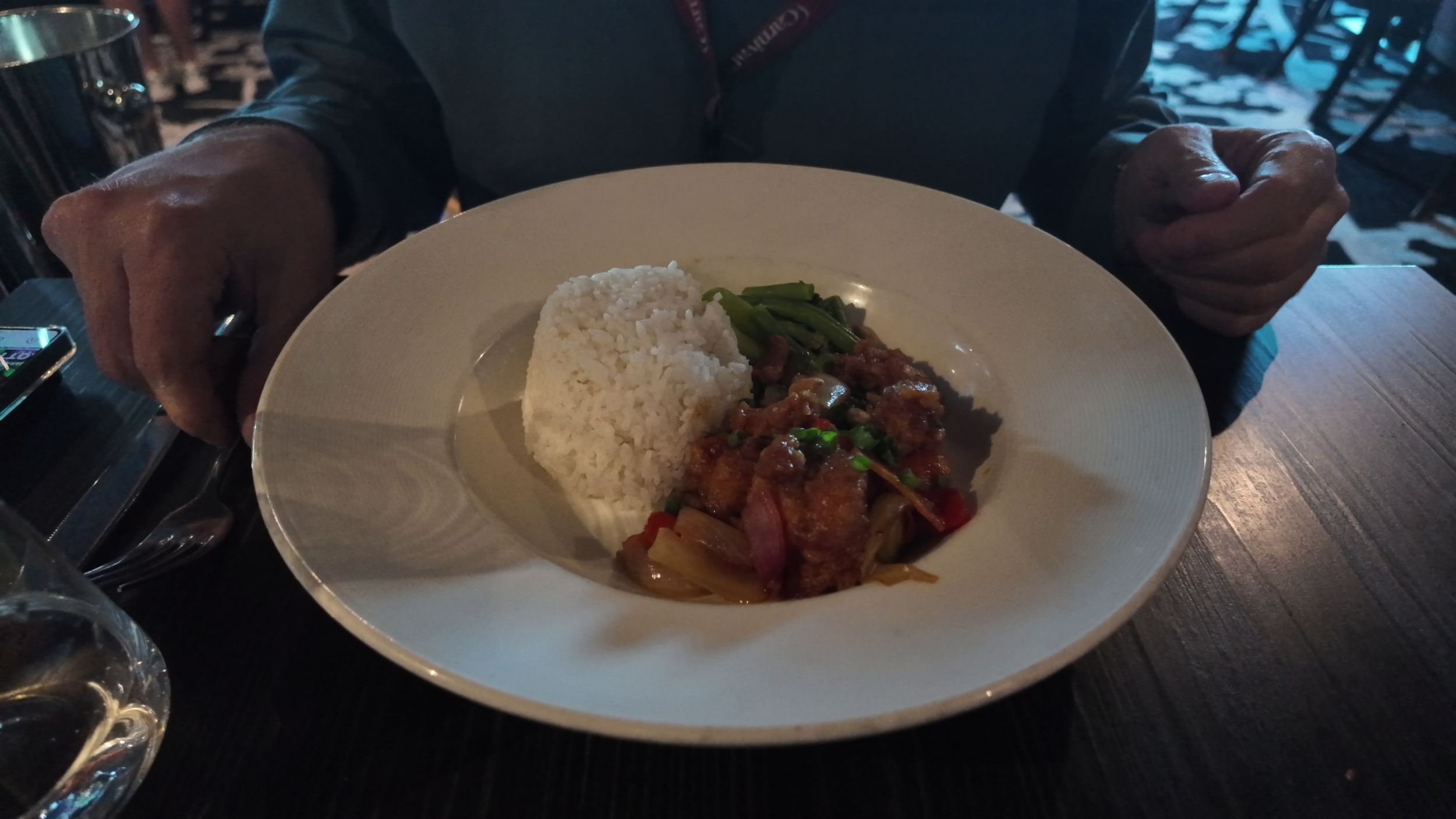 Plate of food with rice, stir-fried vegetables, and a spicy meat dish, on a dark wooden table, with a glass of water nearby.