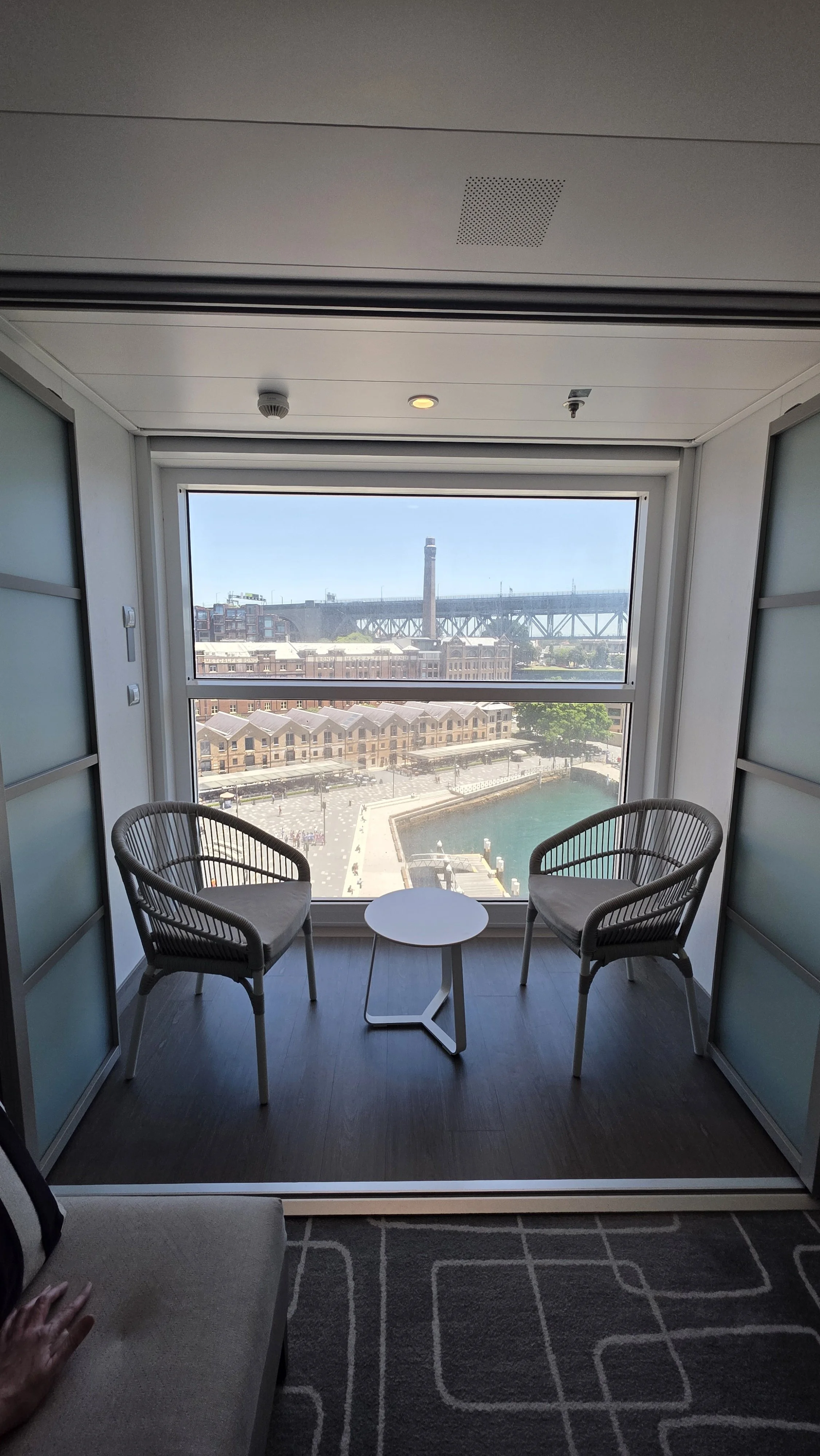 A room with two chairs and a small round table facing a large window with a view of a cityscape, including a bridge and water.