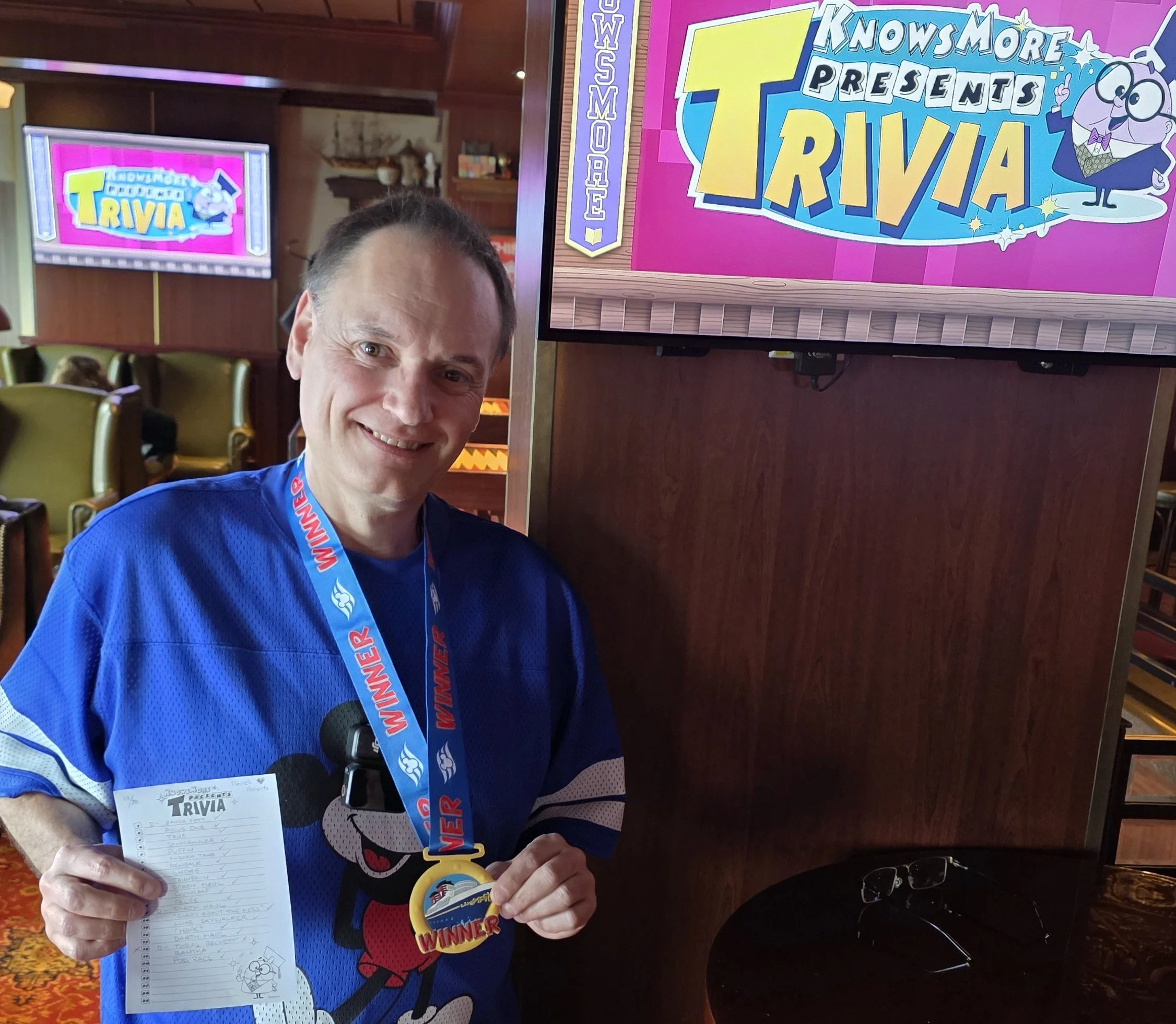 A man smiling, wearing a blue sports jersey and a winner's medal, is holding a sheet of paper with a trivia game score. Behind him is a screen displaying a trivia game with colorful graphics and cartoons, in a room with wooden walls and chairs.