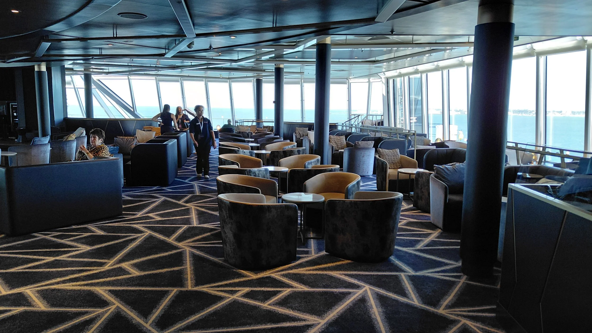 Inside view of an airport lounge with black and beige armchairs, small round tables, large windows showing the sea, and passengers and staff.