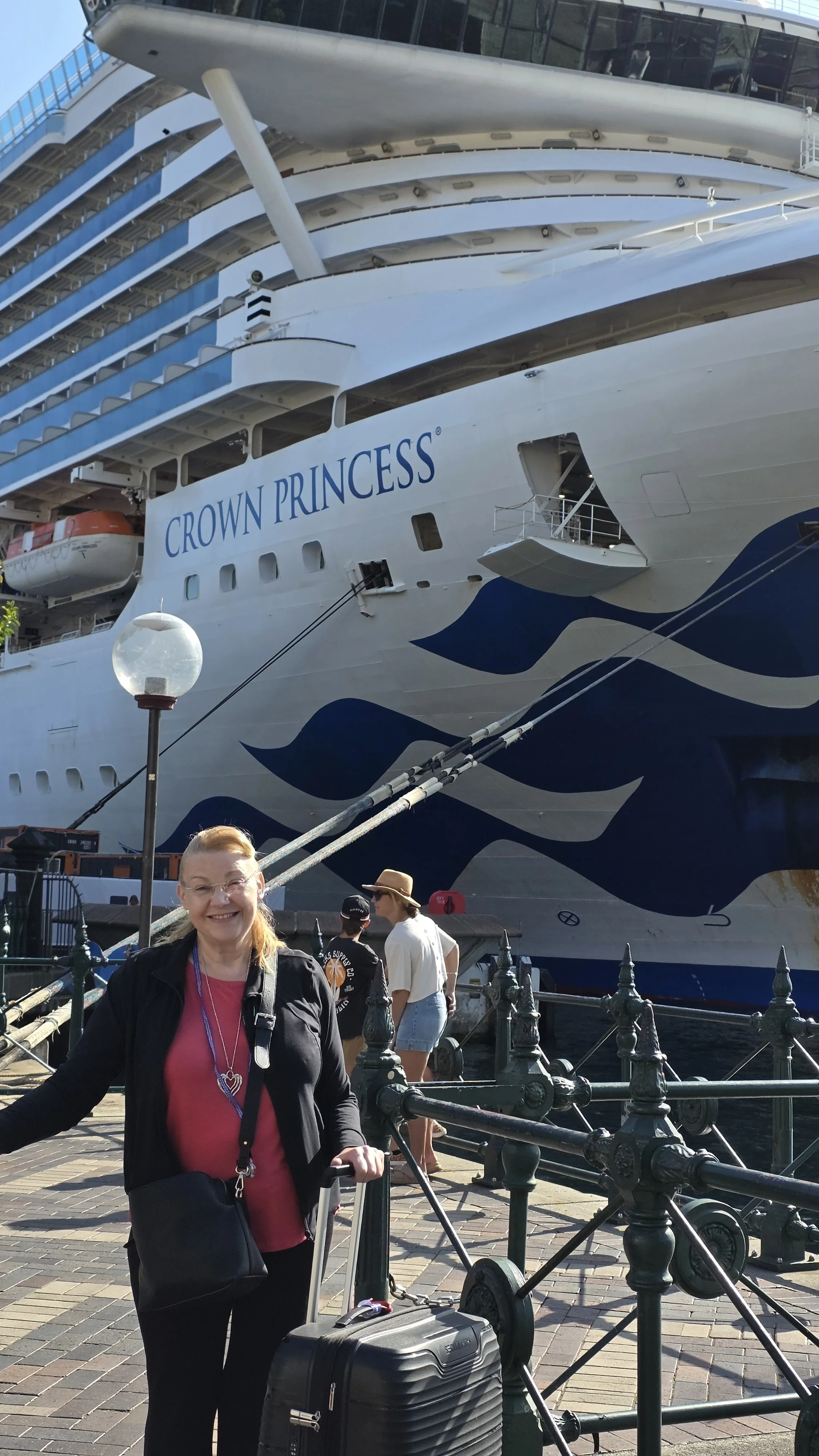Smiling woman standing with a suitcase in front of a large cruise ship named Crown Princess, docked at a port with other people nearby.