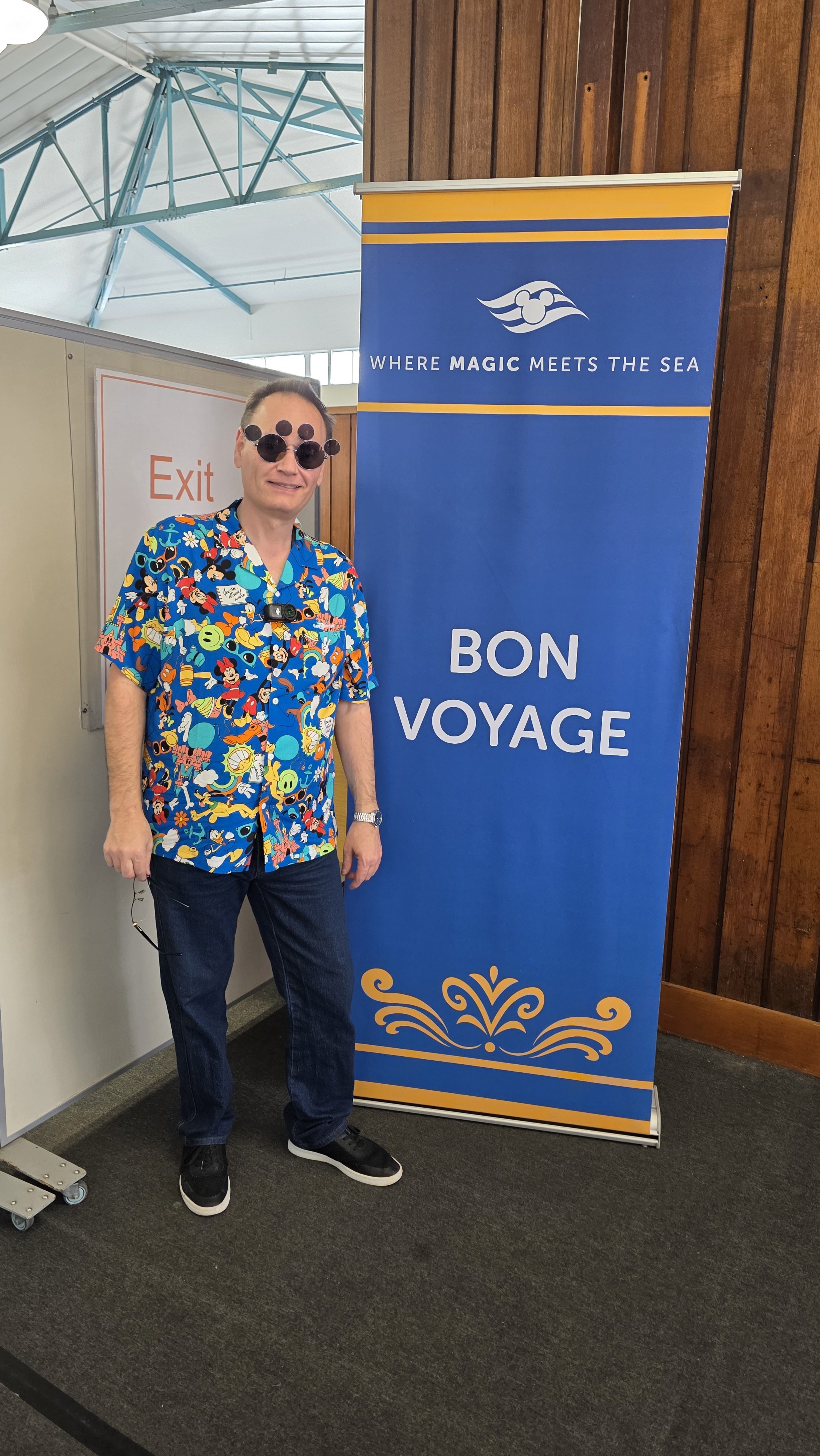 A man standing next to a blue banner that says "Bon Voyage" and "Where magic meets the sea," with a person wearing a colorful Hawaiian shirt, sunglasses, and a camera around his neck.