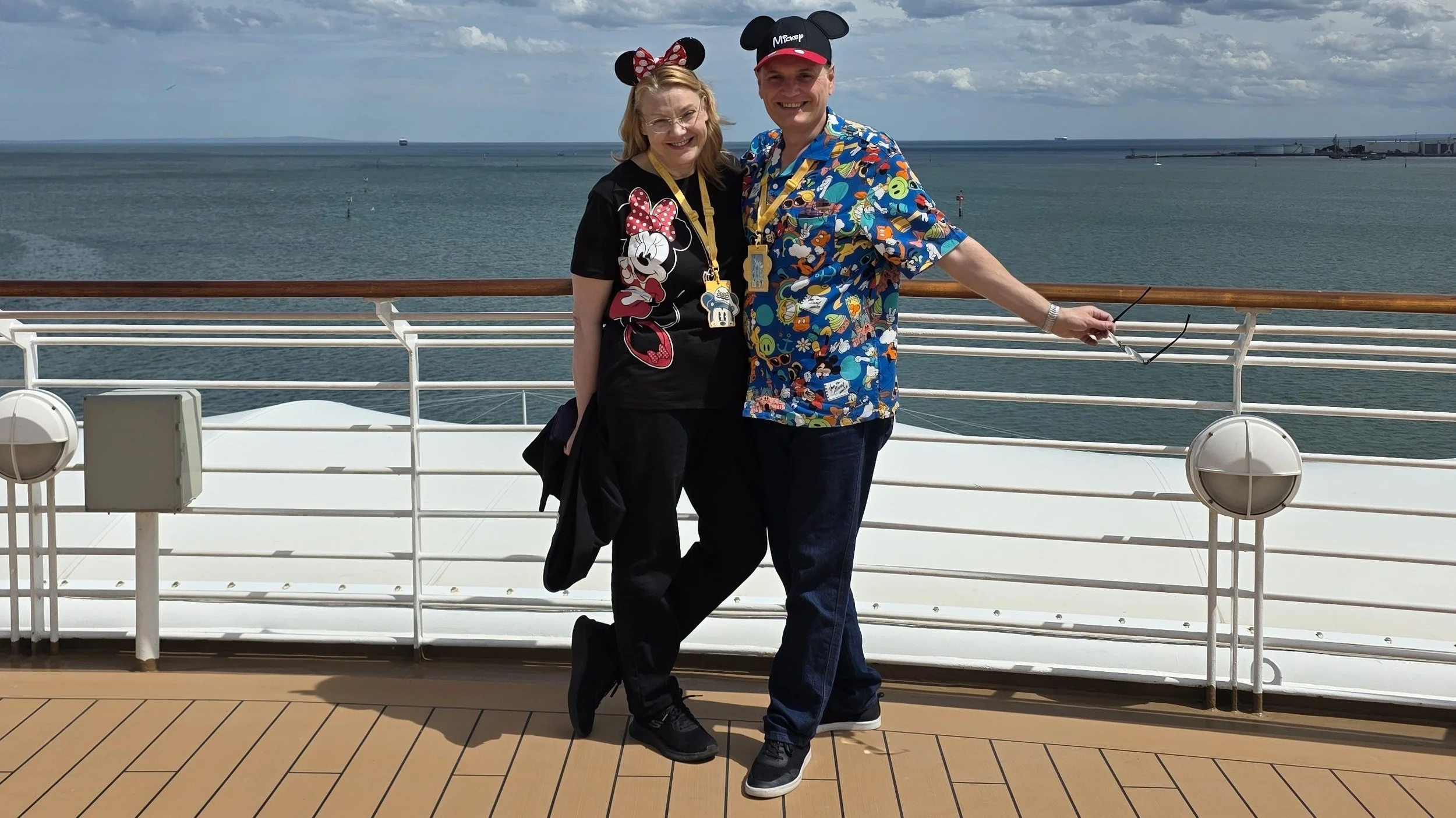 A smiling woman and man dressed in Disney-themed clothing and hats, wearing Disney character ears, pose together on a cruise ship deck with the ocean and cloudy sky in the background.