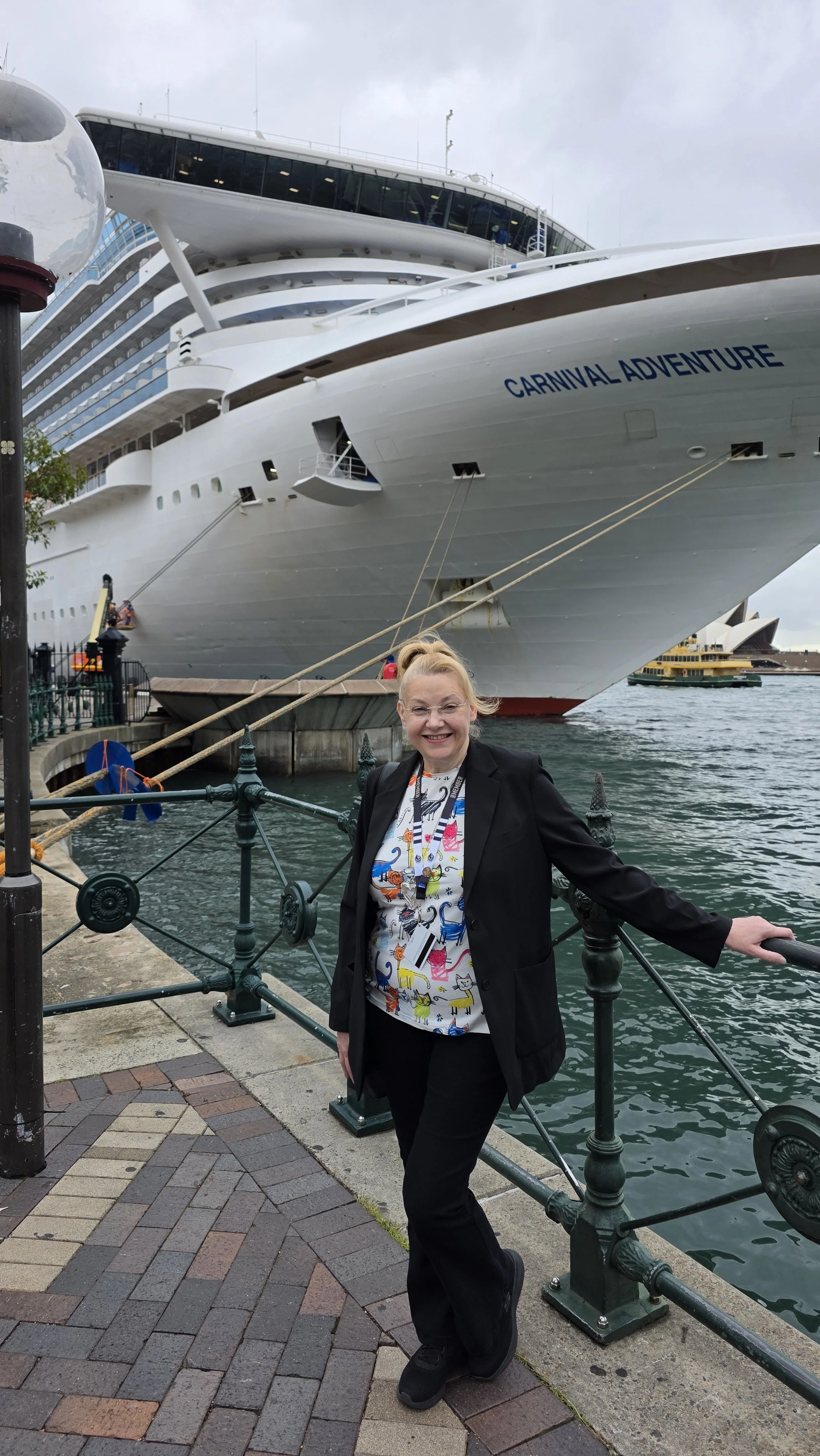 A smiling woman standing on a waterfront promenade with a cruise ship named Carnival Adventure docked behind her.