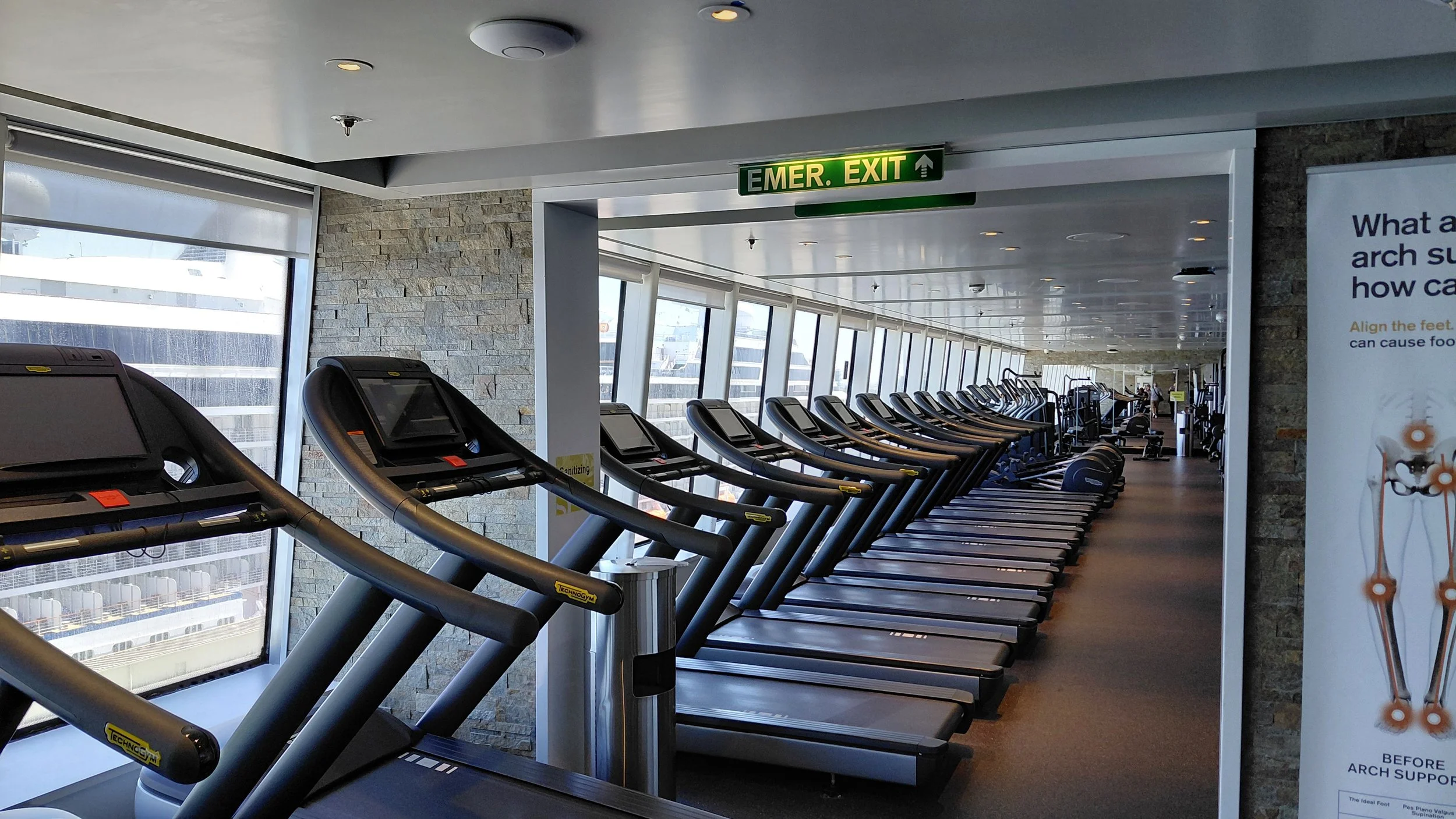 A row of treadmills in a modern gym with large windows and an emergency exit sign overhead.